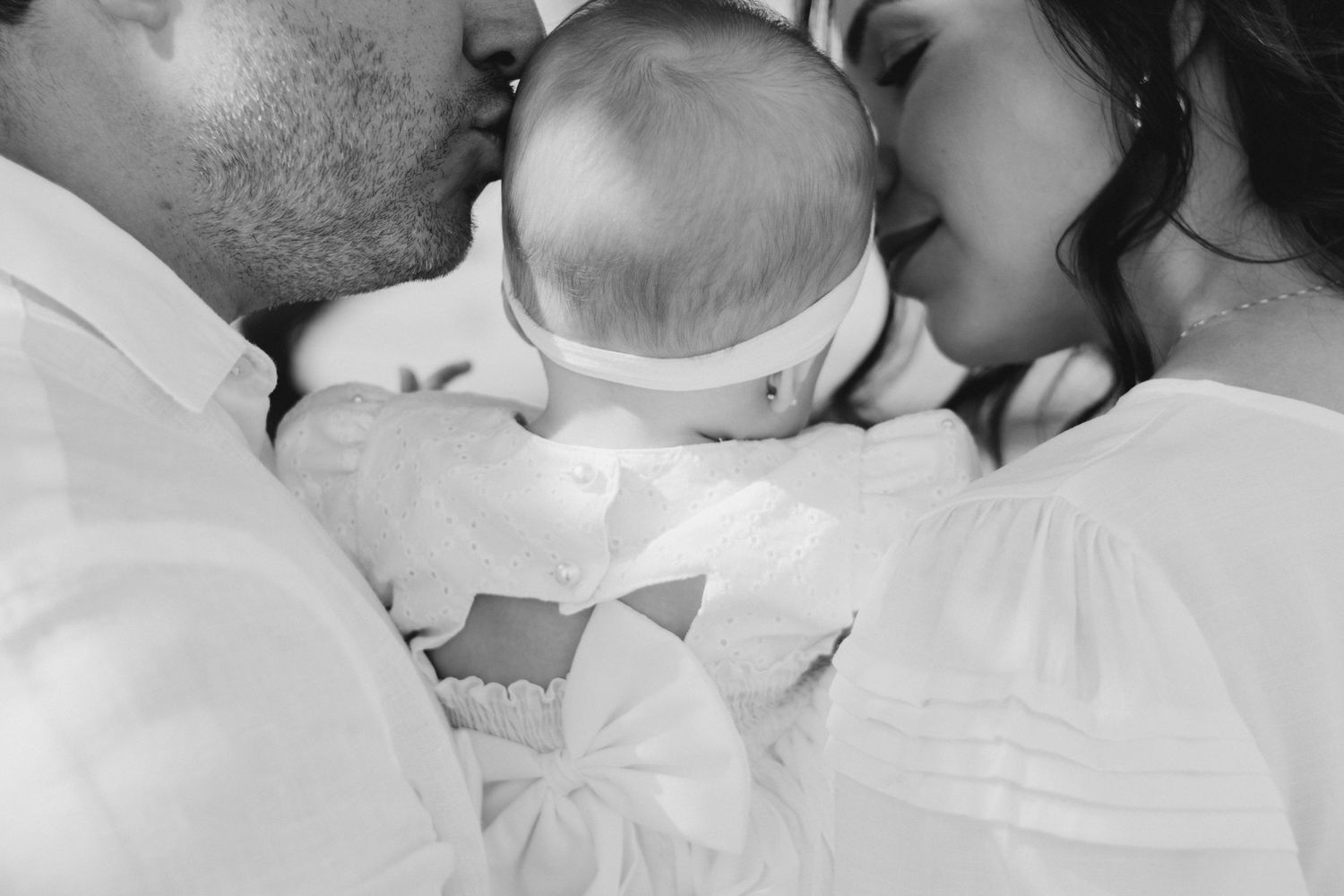 Black and white photo of parents kissing their newborn baby dressed in white.