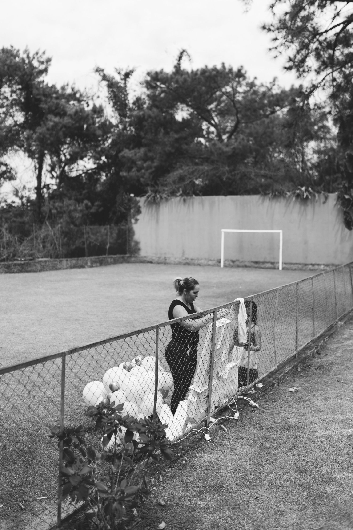 Black and white photo of a woman leaning on a chain-link fence beside a soccer field with a goal post.