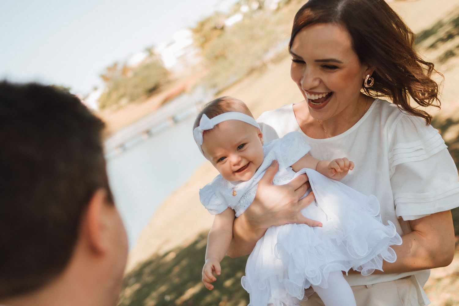 A joyful mother in white holds a smiling baby girl with a bow headband outdoors near water.
