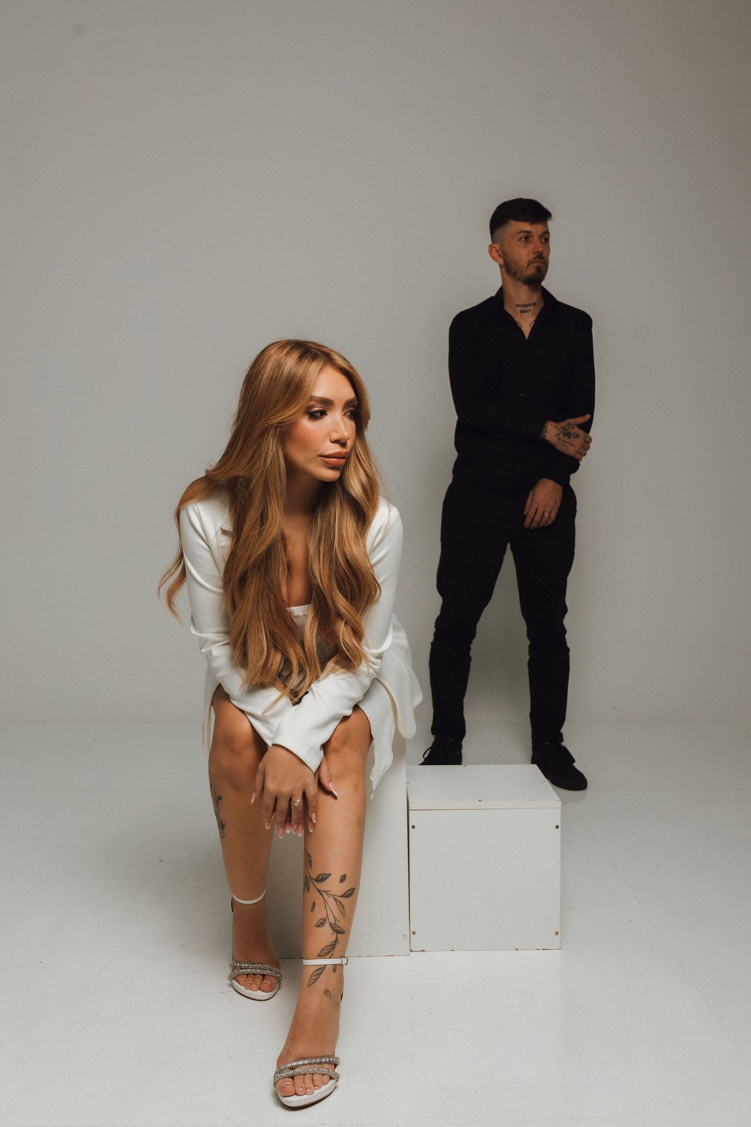 A woman in white sits on a cube while a tattooed man in black stands behind her in a studio.