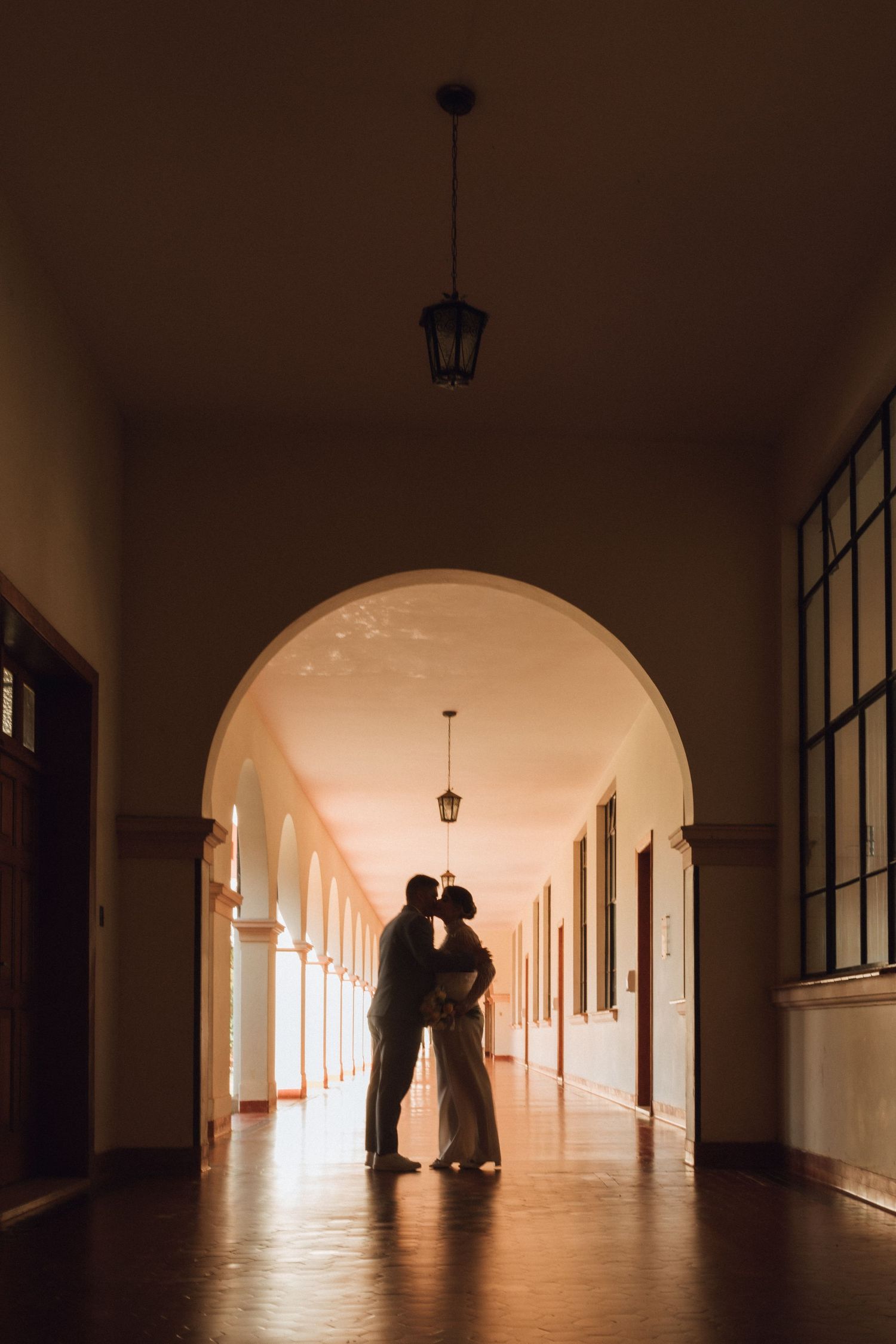 Romantic couple silhouette kissing in a sunlit arched corridor with warm tones and hanging lanterns.
