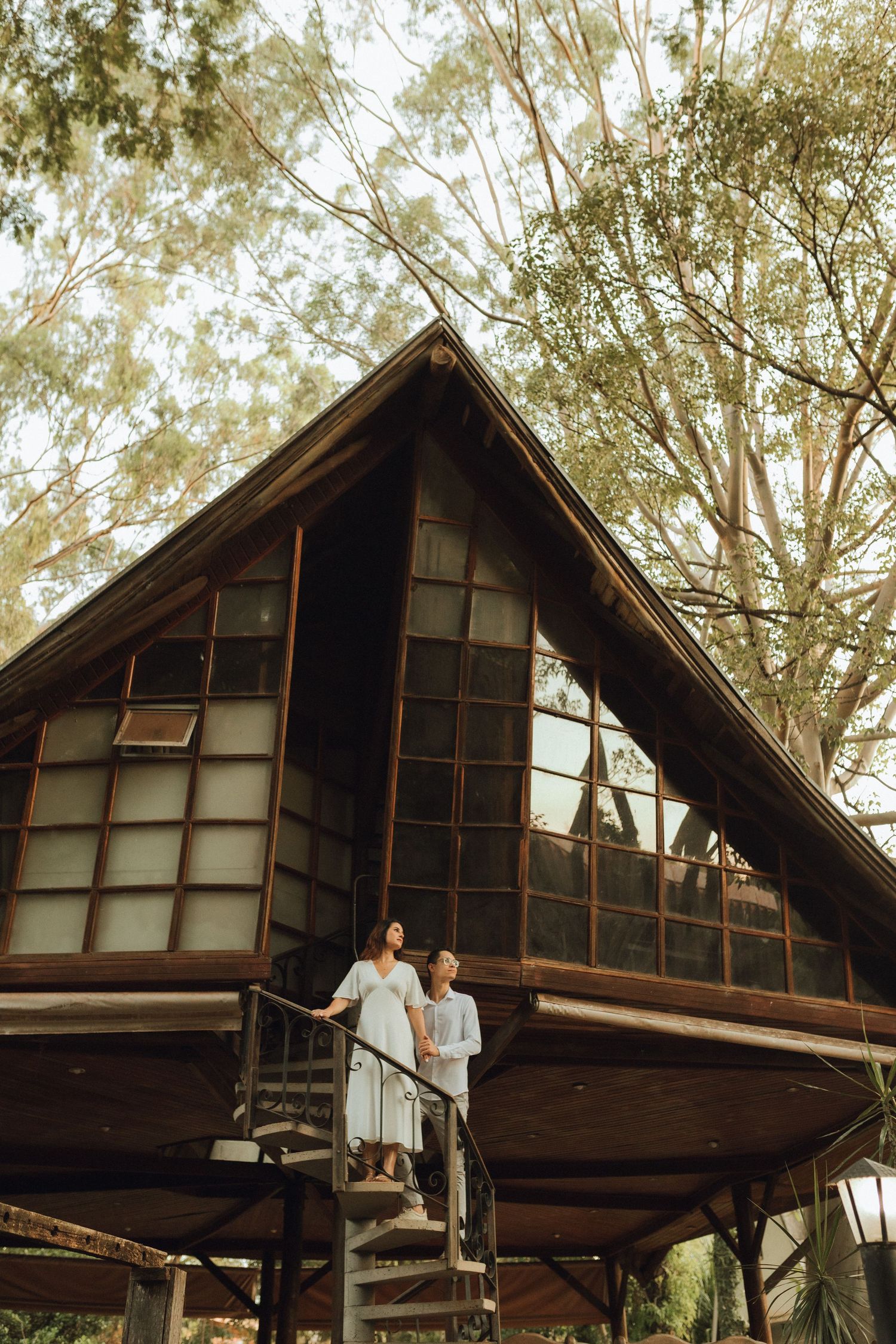 Couple standing on spiral staircase of a rustic wooden A-frame cabin surrounded by tall eucalyptus trees.