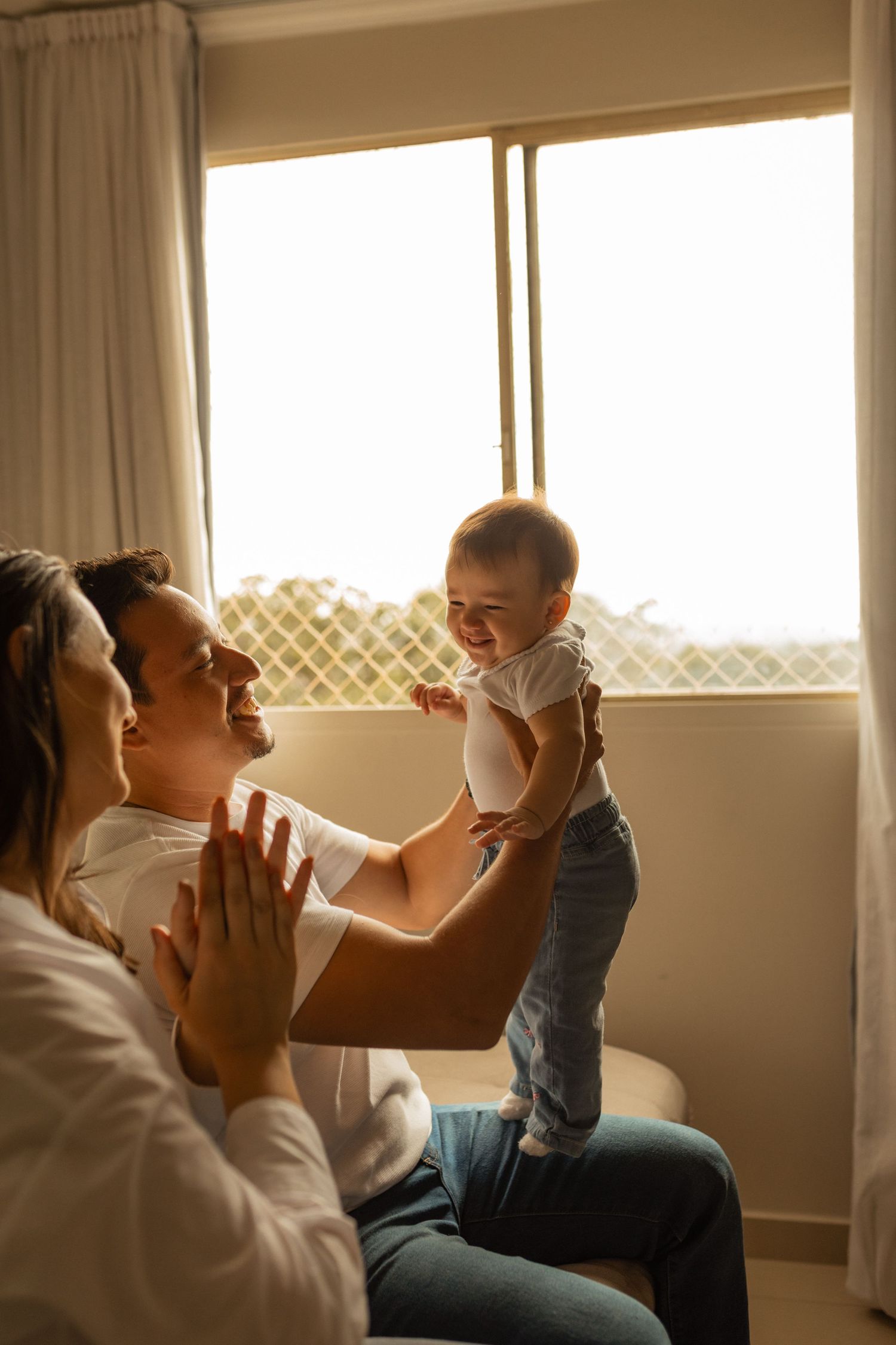 Happy parents playing with smiling baby near sunny window at home.