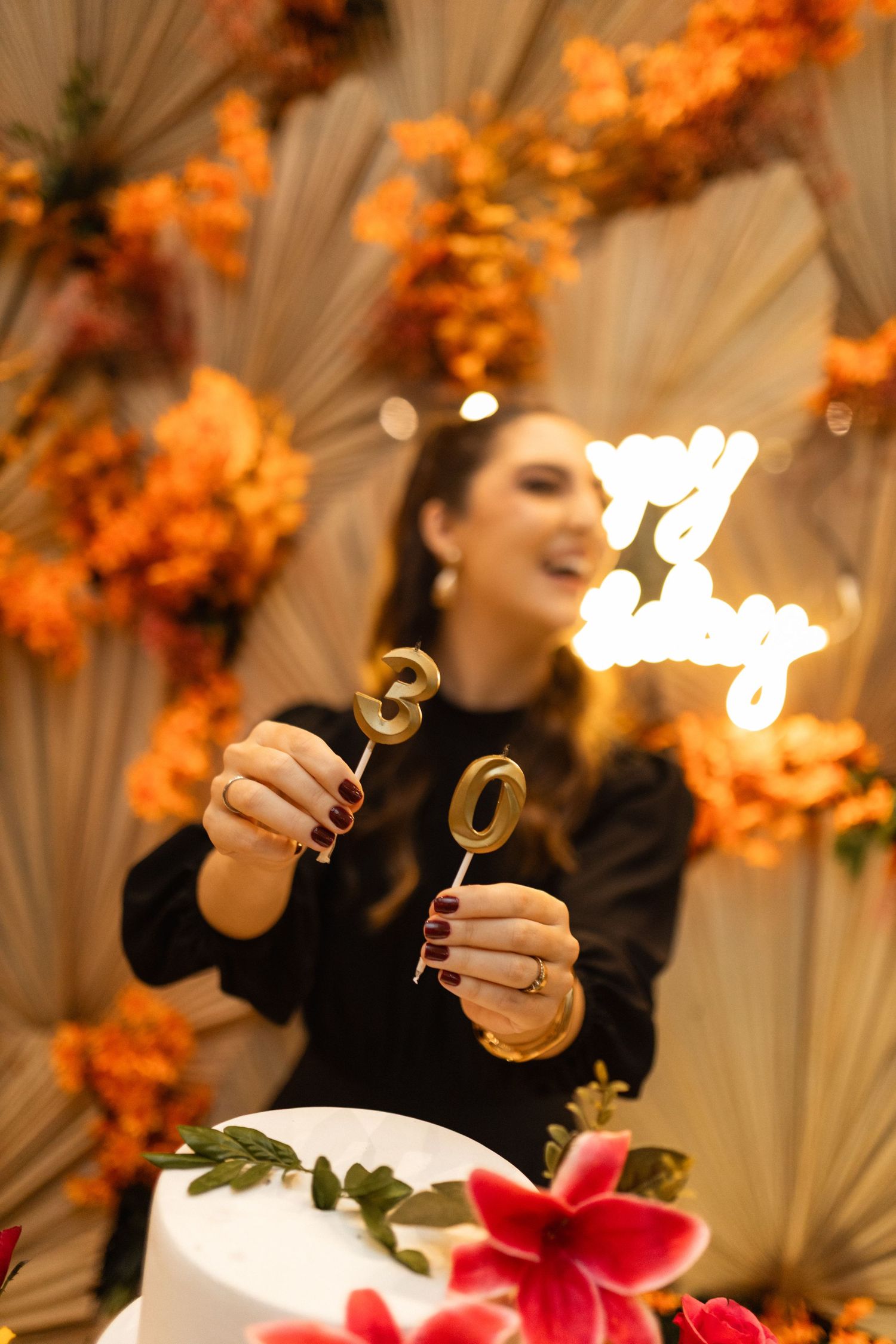 Woman holding gold number 30 cake toppers at birthday party with orange floral decoration behind.