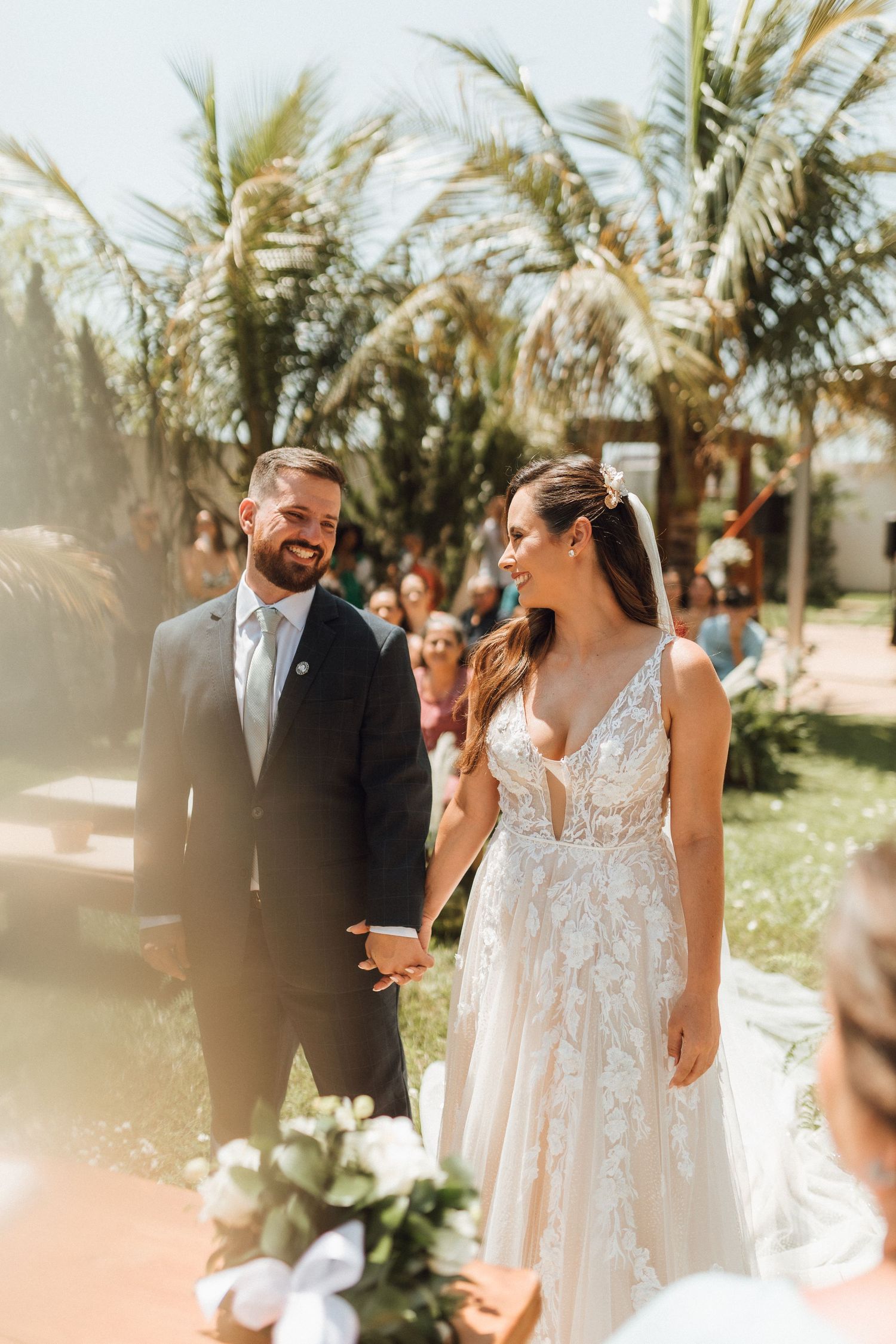 Happy bride and groom holding hands during outdoor wedding ceremony surrounded by palm trees and guests.