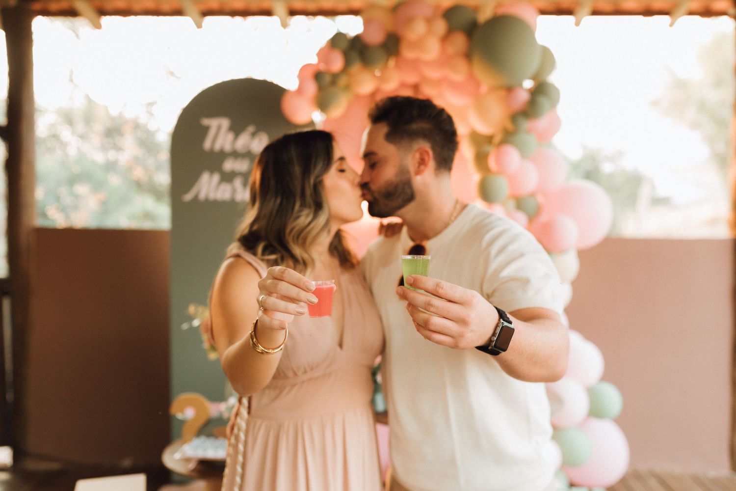 Couple sharing a romantic kiss at a gender reveal party with pink and green balloon arch backdrop.