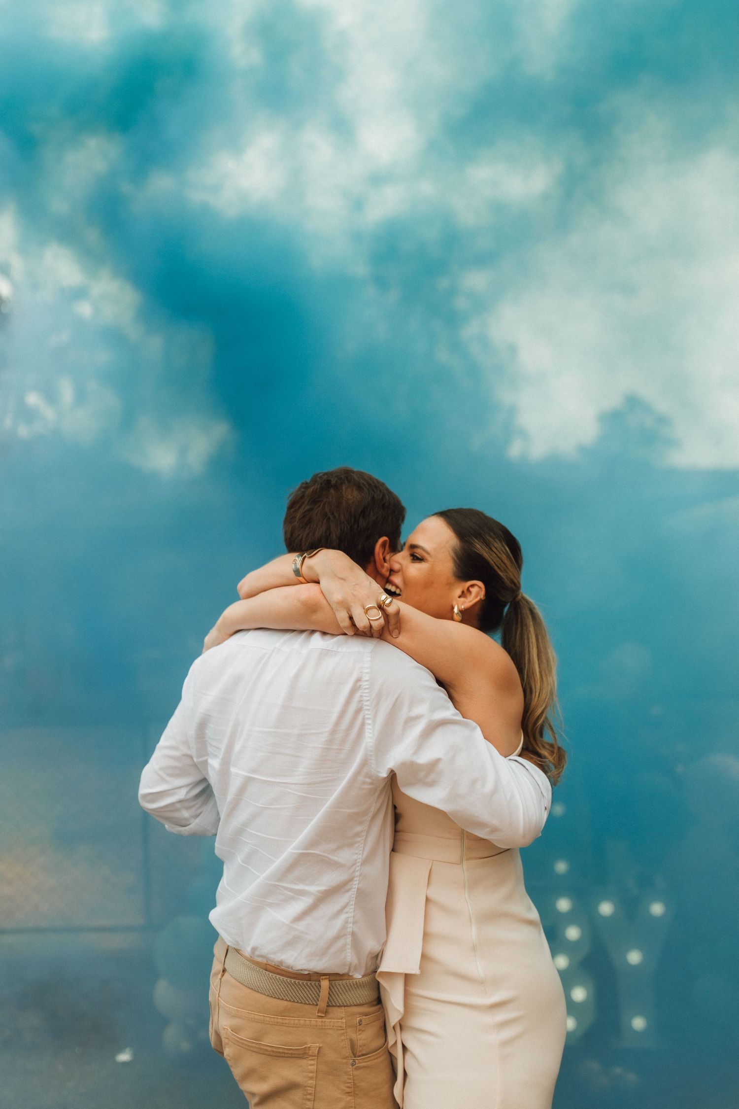 Couple embracing and kissing against a dramatic blue smoke cloud backdrop at a gender reveal celebration.