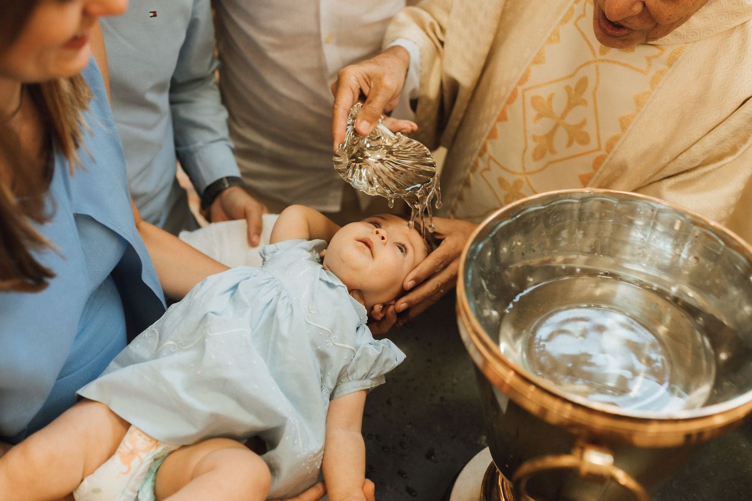 Priest performing Catholic infant baptism ceremony, pouring holy water over baby's head at baptismal font.