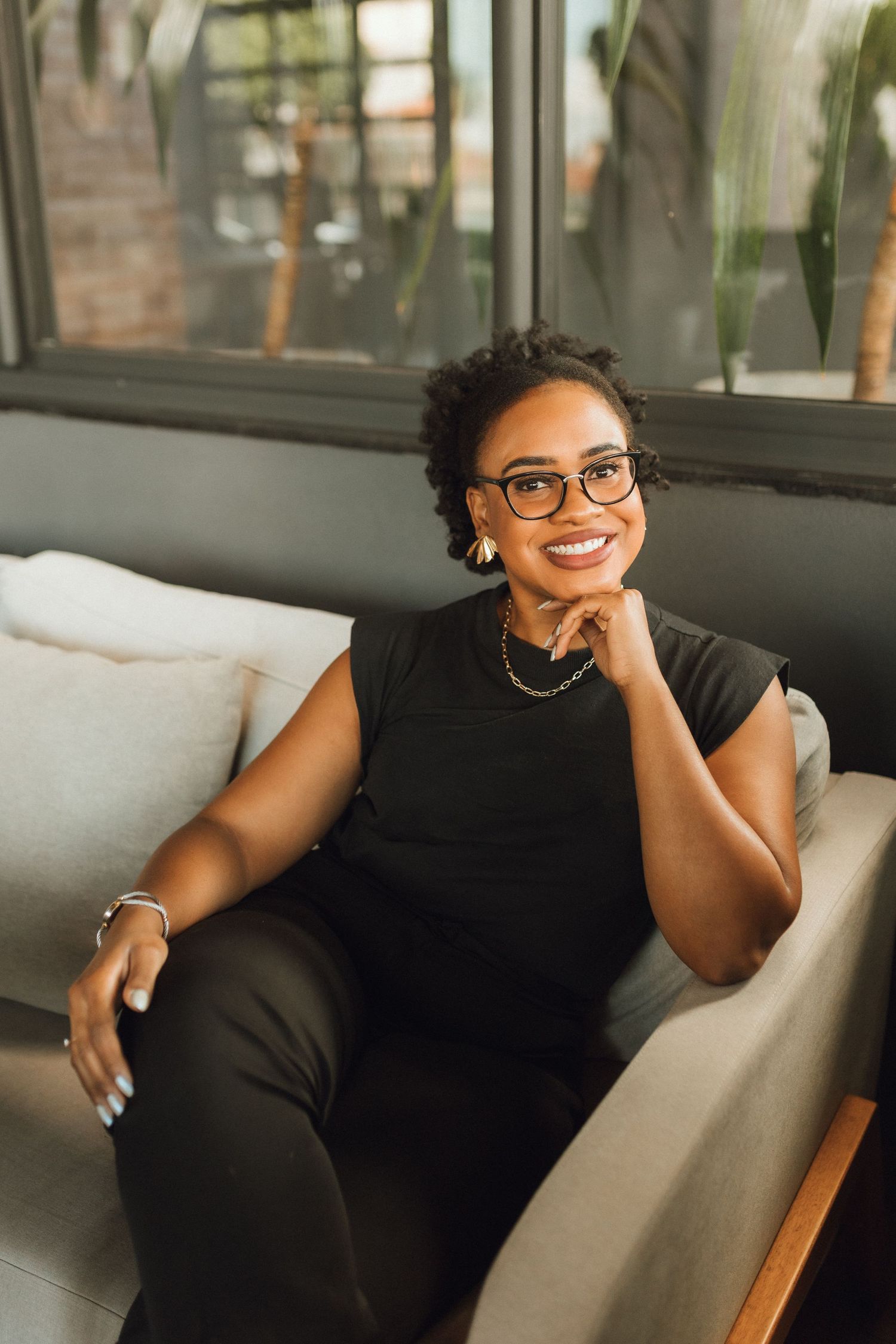 Smiling Black woman in glasses and black outfit relaxing on gray sofa indoors.