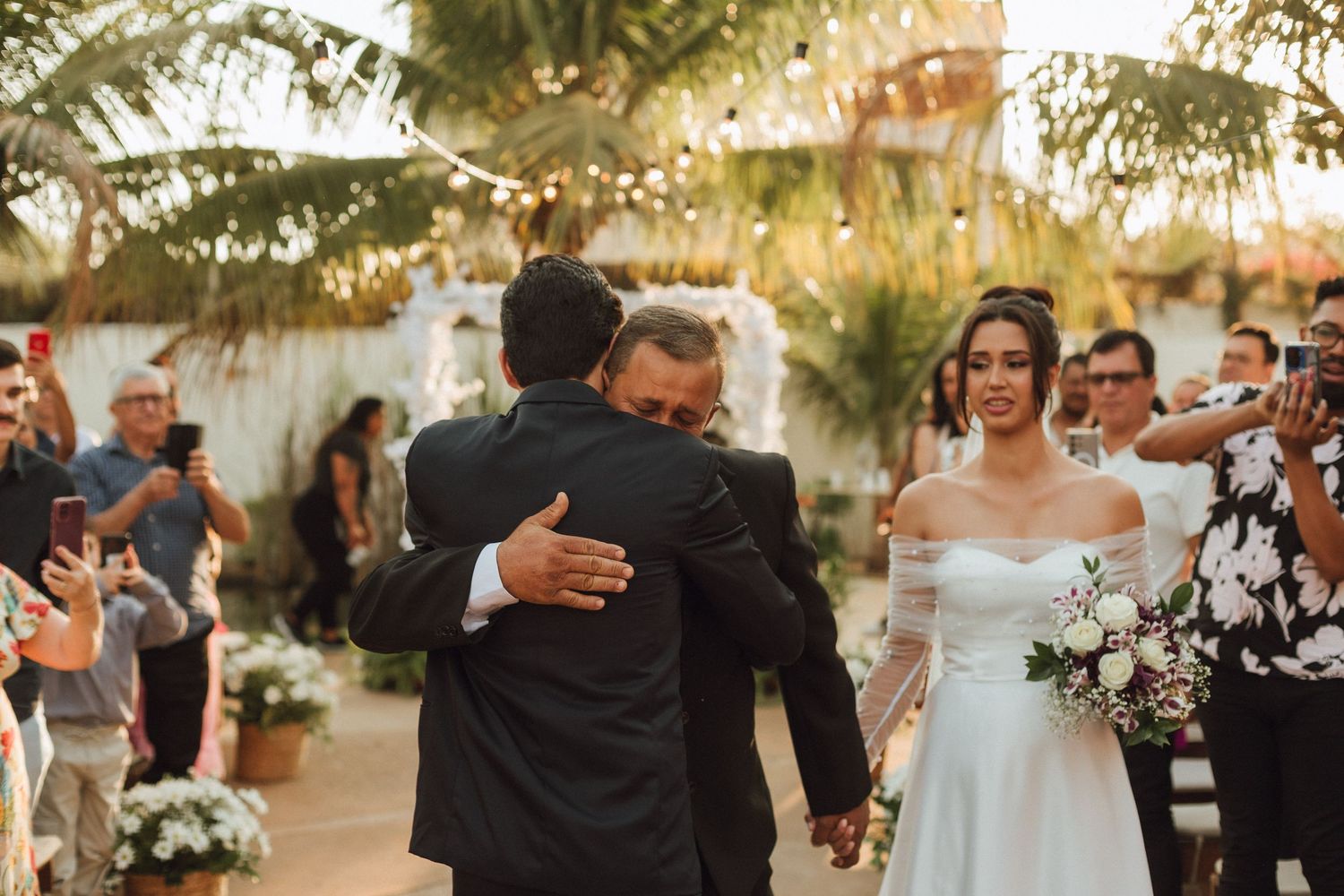 Groom embracing father-in-law while bride holds bouquet during outdoor wedding ceremony.