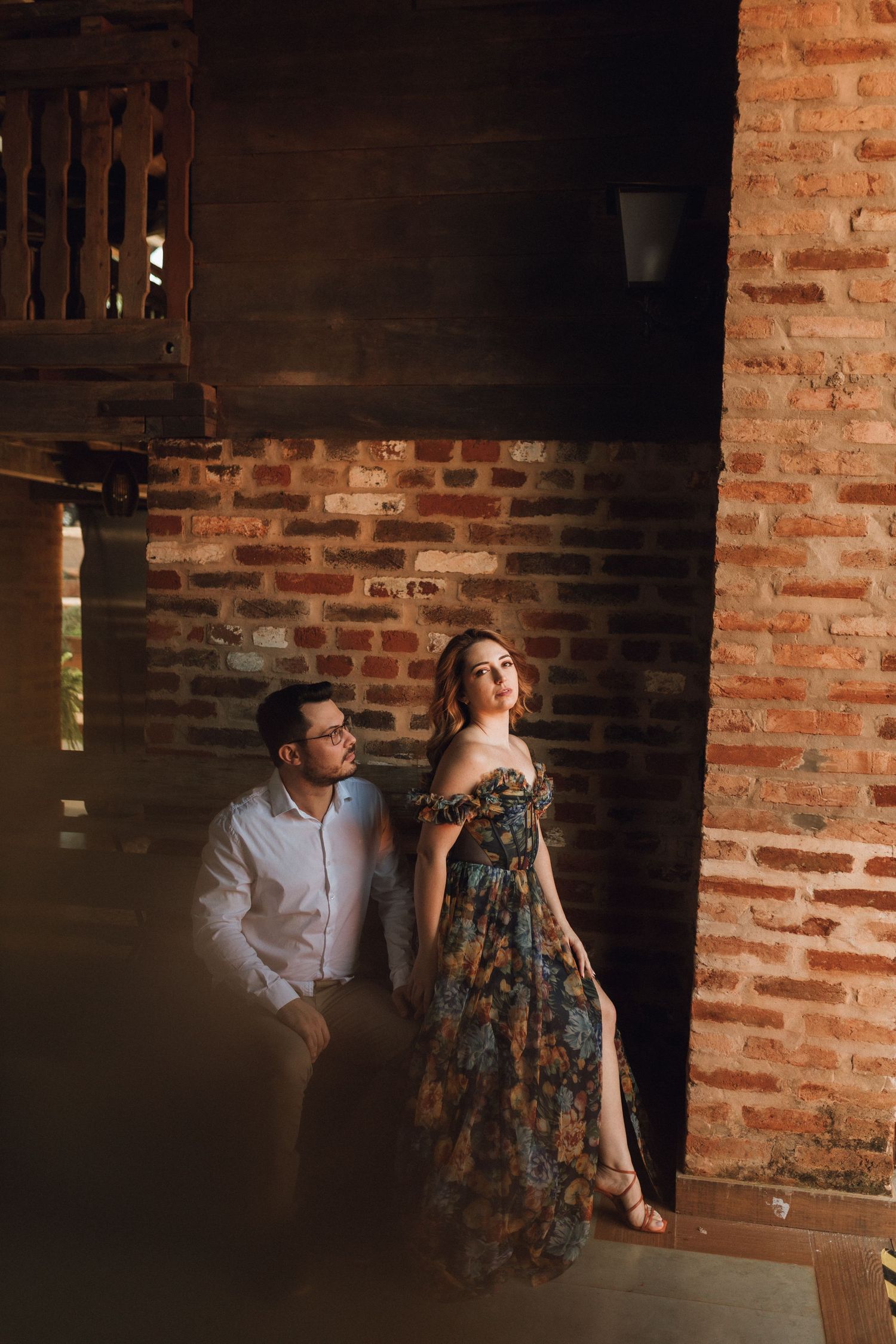 Elegant couple posing against a rustic brick wall, woman in floral dress, man in white shirt.