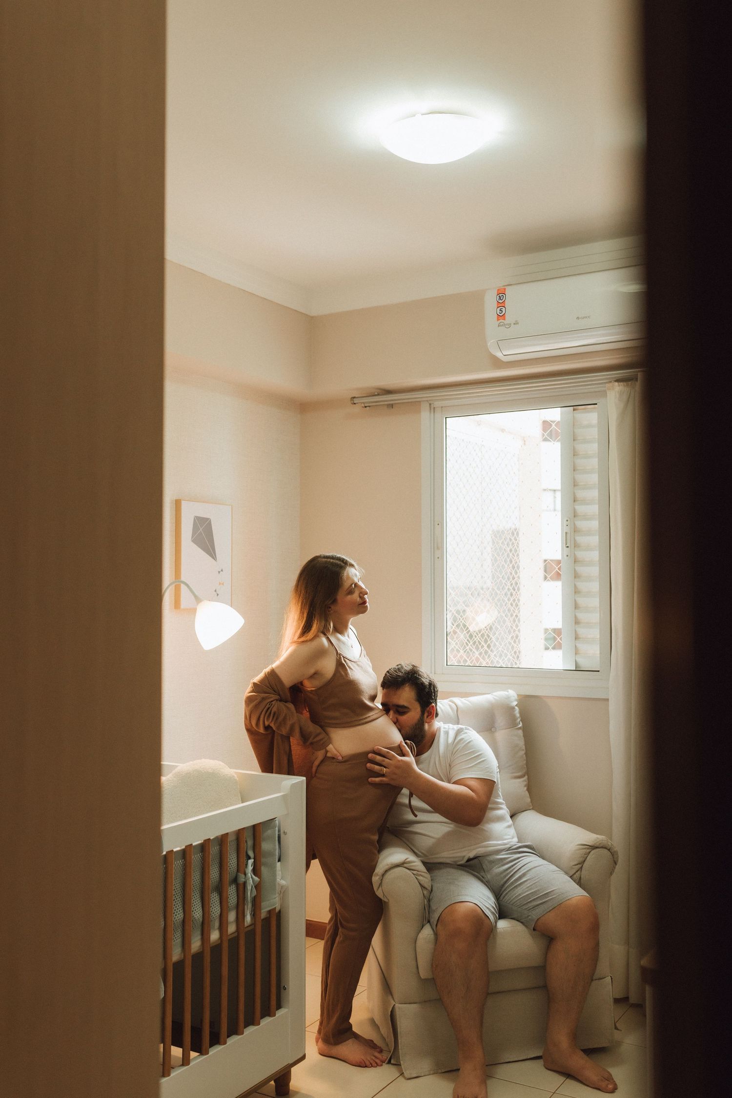 Man seated in nursery chair gently touches pregnant partners belly near baby crib.
