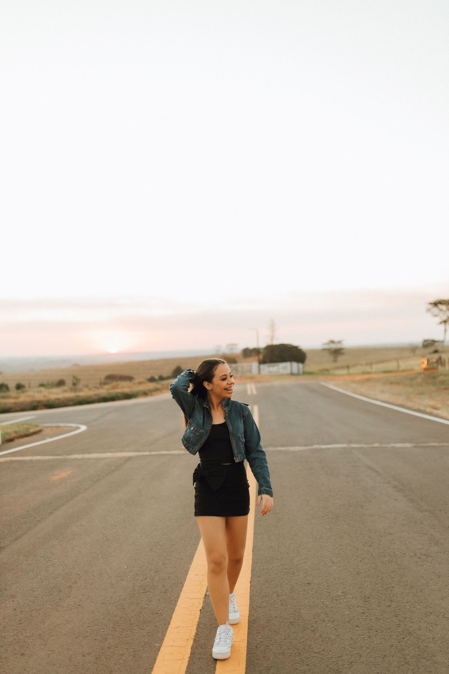A smiling young woman in a black mini dress, denim jacket, and white sneakers walks along a road at sunset.