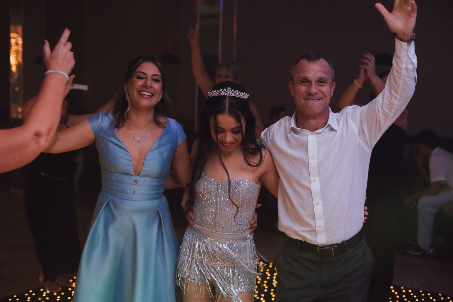 Three people smile and celebrate together at quinceañera party with tiara-wearing birthday girl in center.