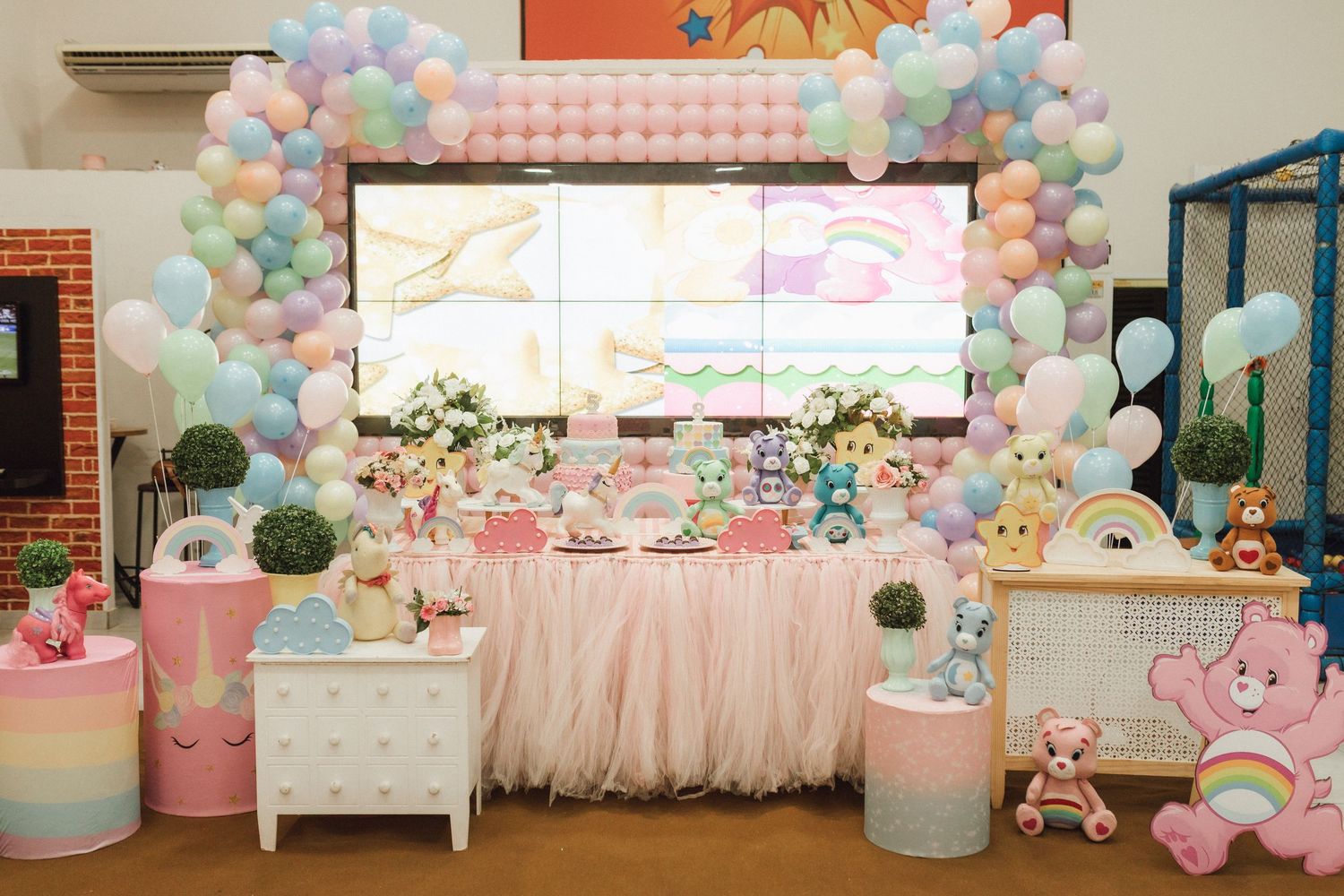 Care Bears themed birthday party decoration with pastel balloons arch, tulle table skirt, and colorful figurines.