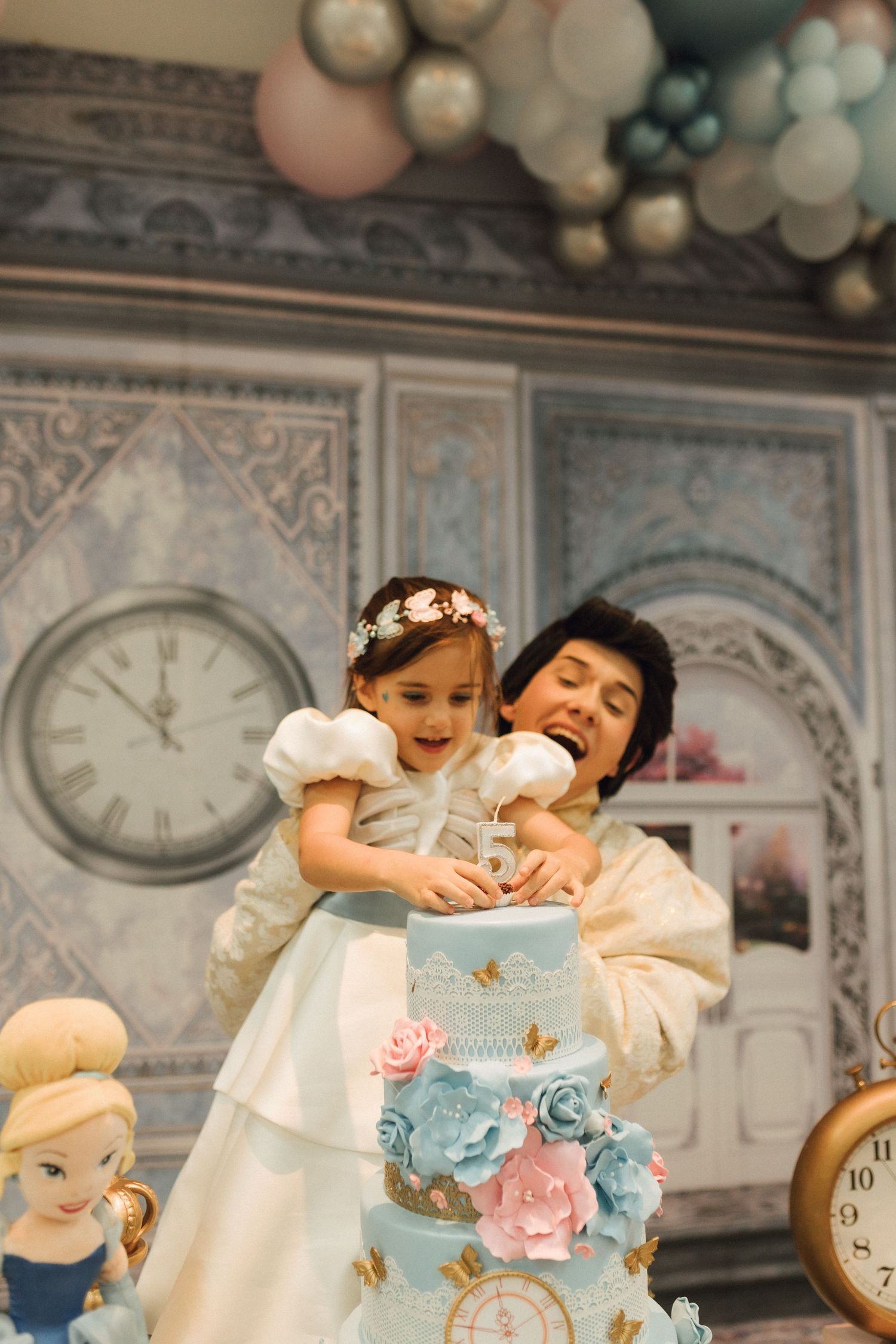 A mother helps her little girl in a white dress cut a blue floral tiered birthday cake at a Cinderella-themed party.