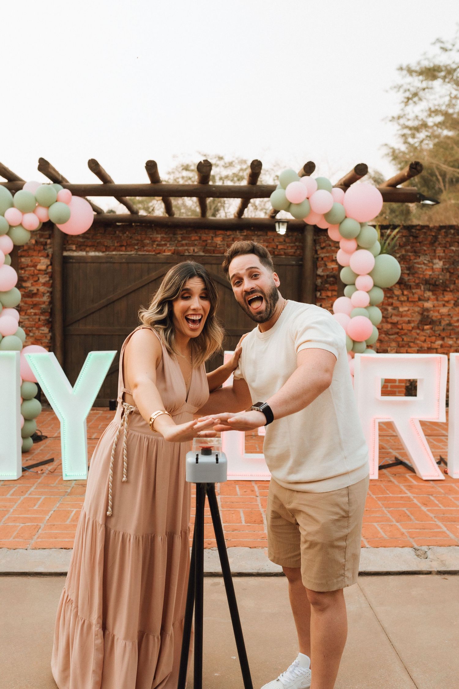 Excited couple posing together at a gender reveal party with pink and green balloon arch and decorative letters.