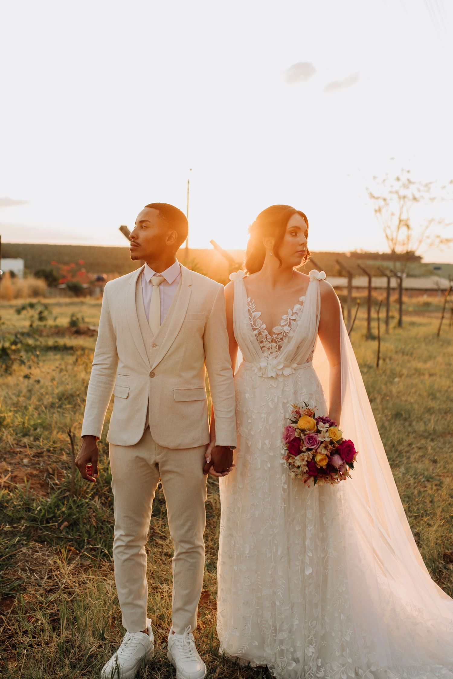 Interracial wedding couple posing elegantly at golden hour, bride holding colorful bouquet in lace gown.
