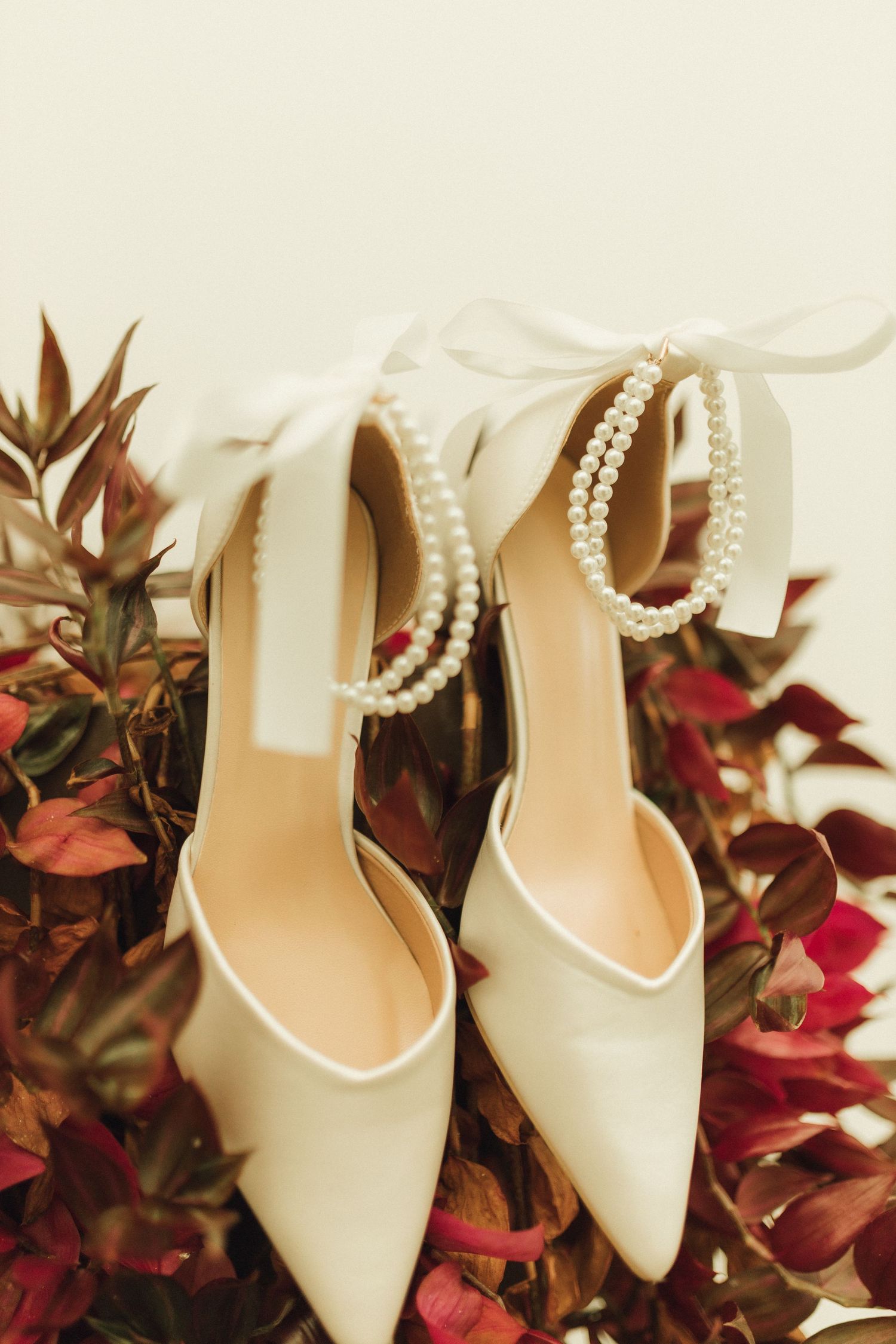 White bridal heels with pearl ankle straps displayed among red and green foliage.