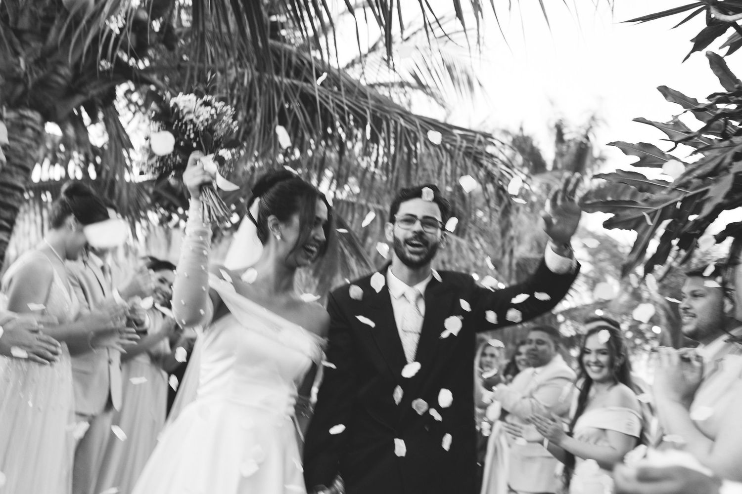 Black and white photo of newlyweds walking through confetti shower after outdoor ceremony.