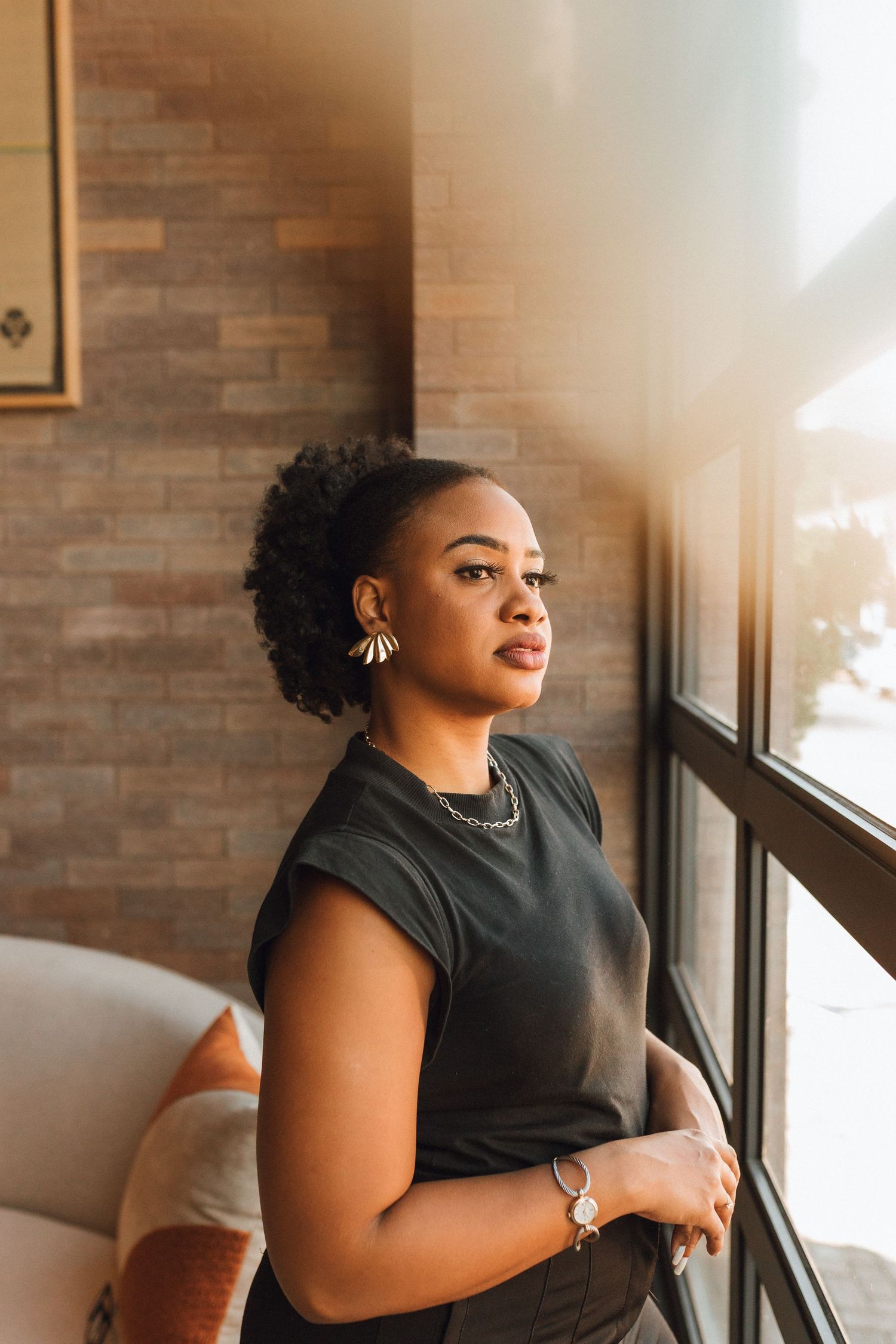 Confident Black woman in black top leaning against brick wall by window in sunlight.
