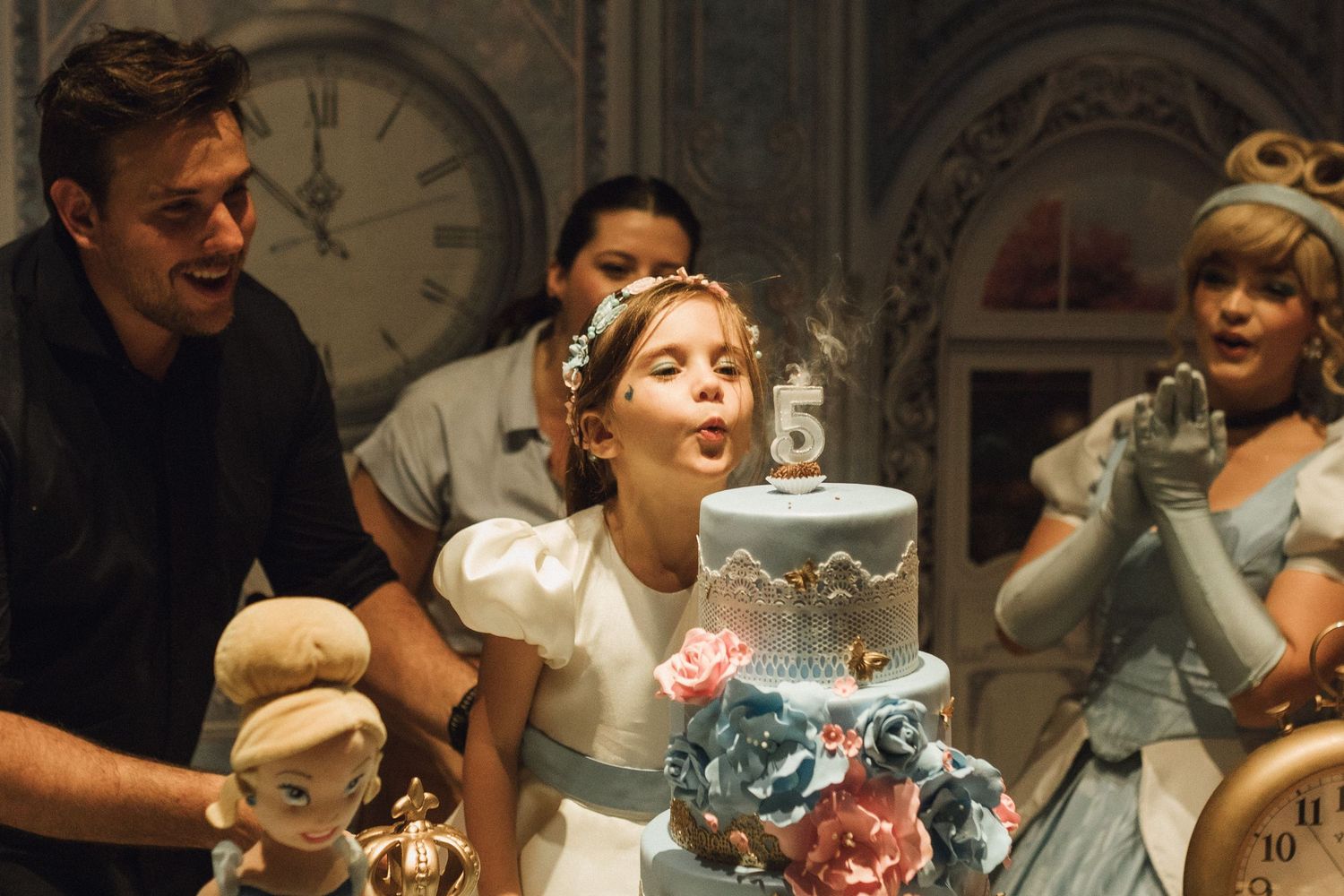 A young girl blows out candles on an elegant tiered birthday cake surrounded by a man and a Cinderella princess character.