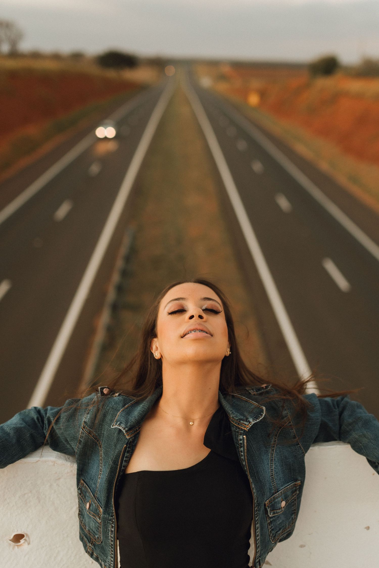 Woman in denim jacket leaning back on road barrier with long straight highway stretching into distance.