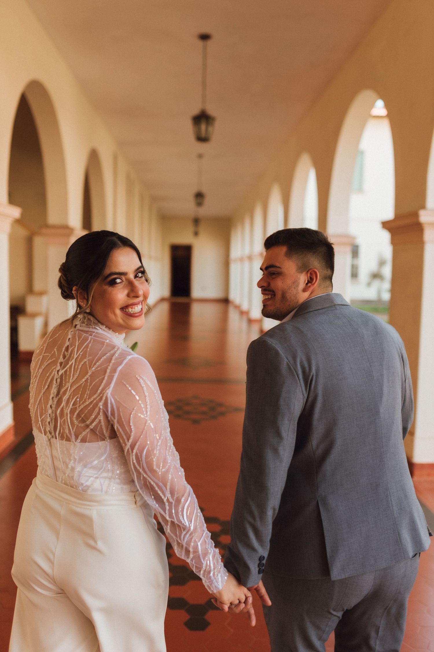Bride and groom walking hand in hand through elegant arched corridor, bride glancing back with a smile on their wedding day.