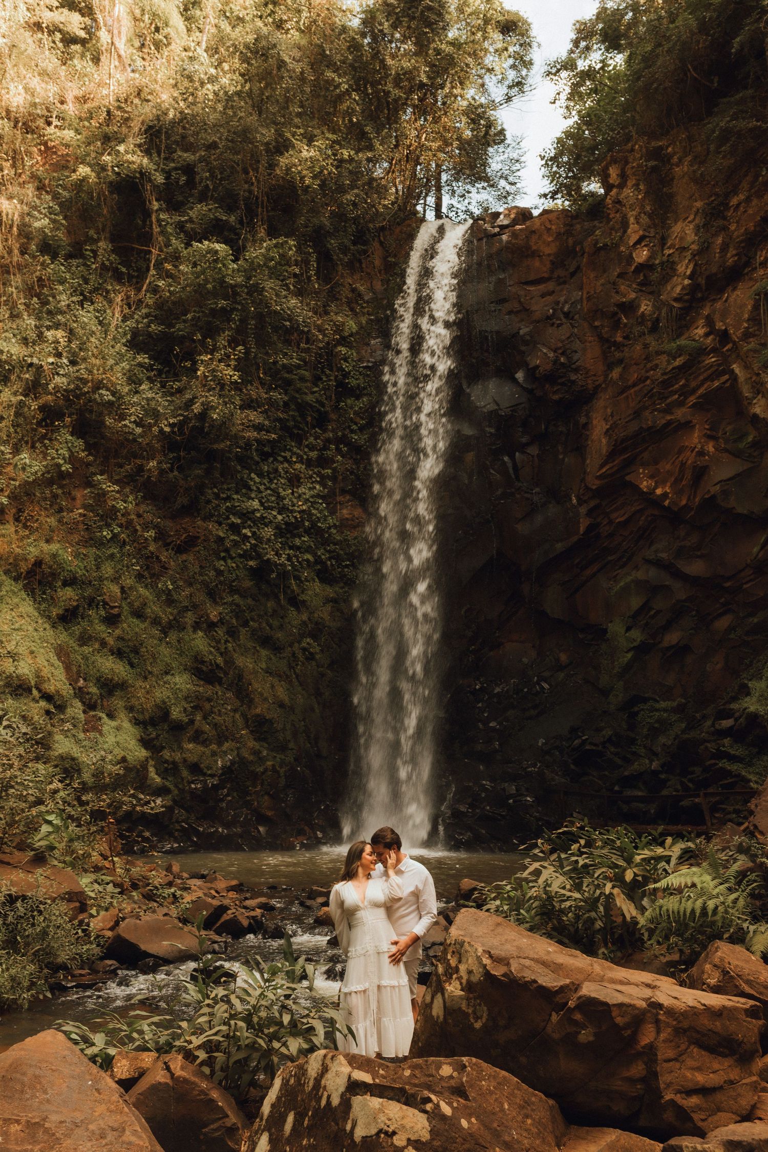 Couple embracing in white outfits before a stunning waterfall surrounded by lush greenery and rocky terrain.