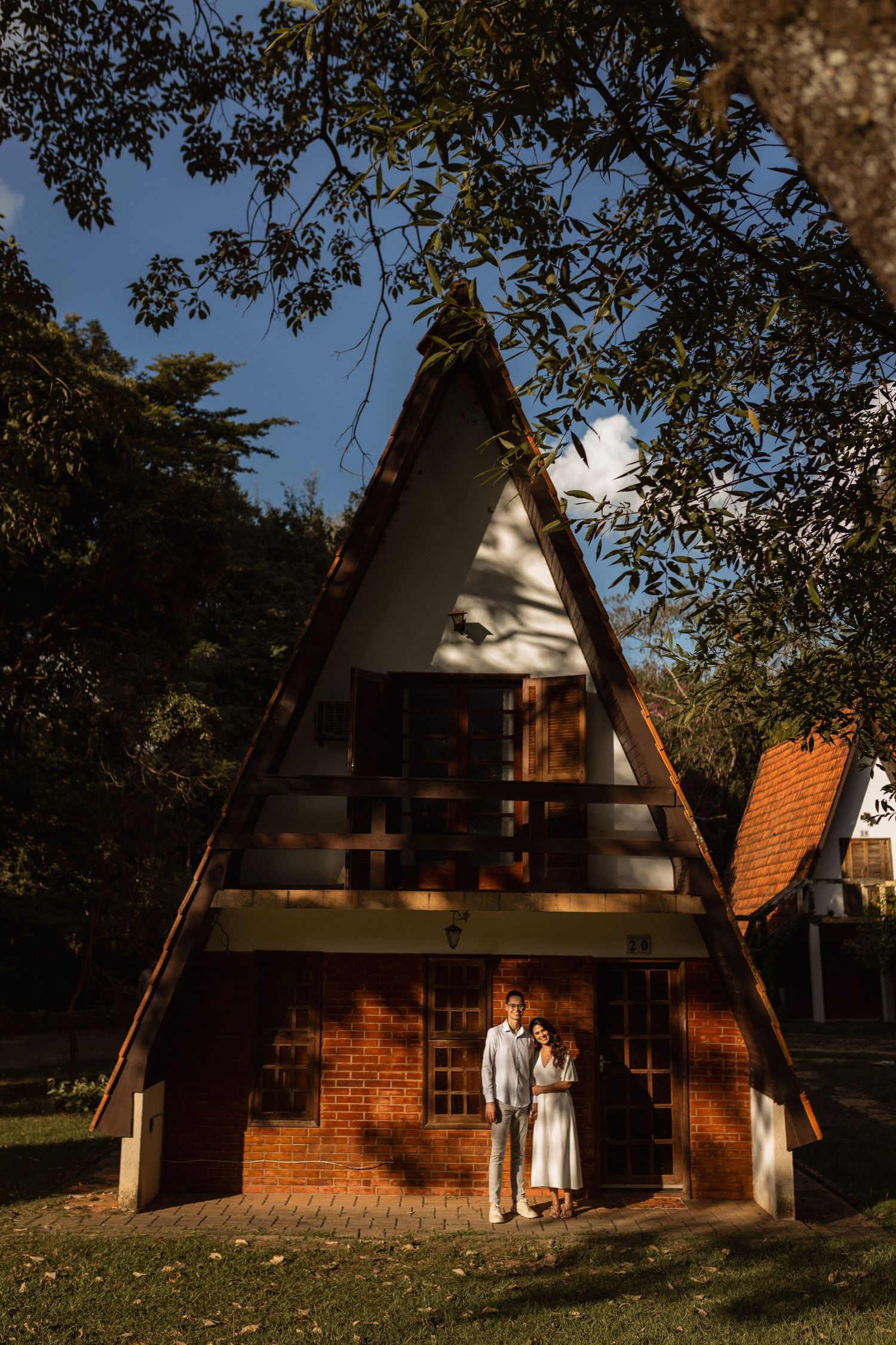 Couple standing in front of a charming A-frame brick cabin surrounded by lush trees on a sunny day.
