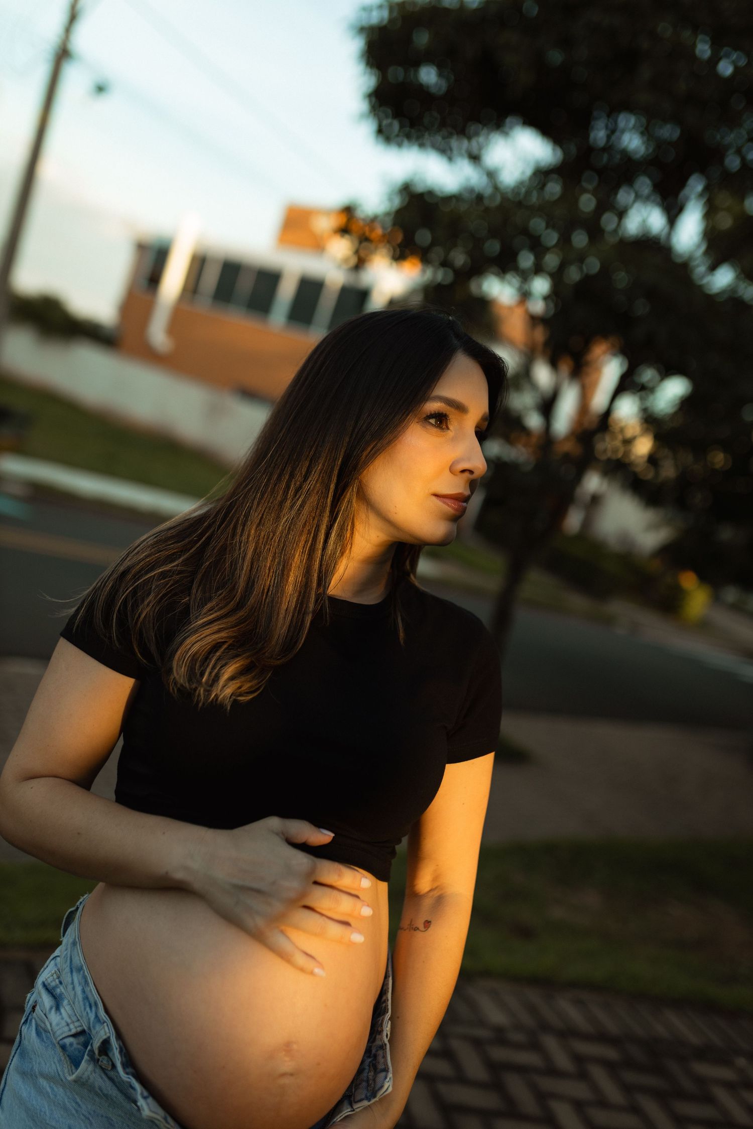 Pregnant woman in black crop top holding baby bump outdoors in warm golden sunset light on street.