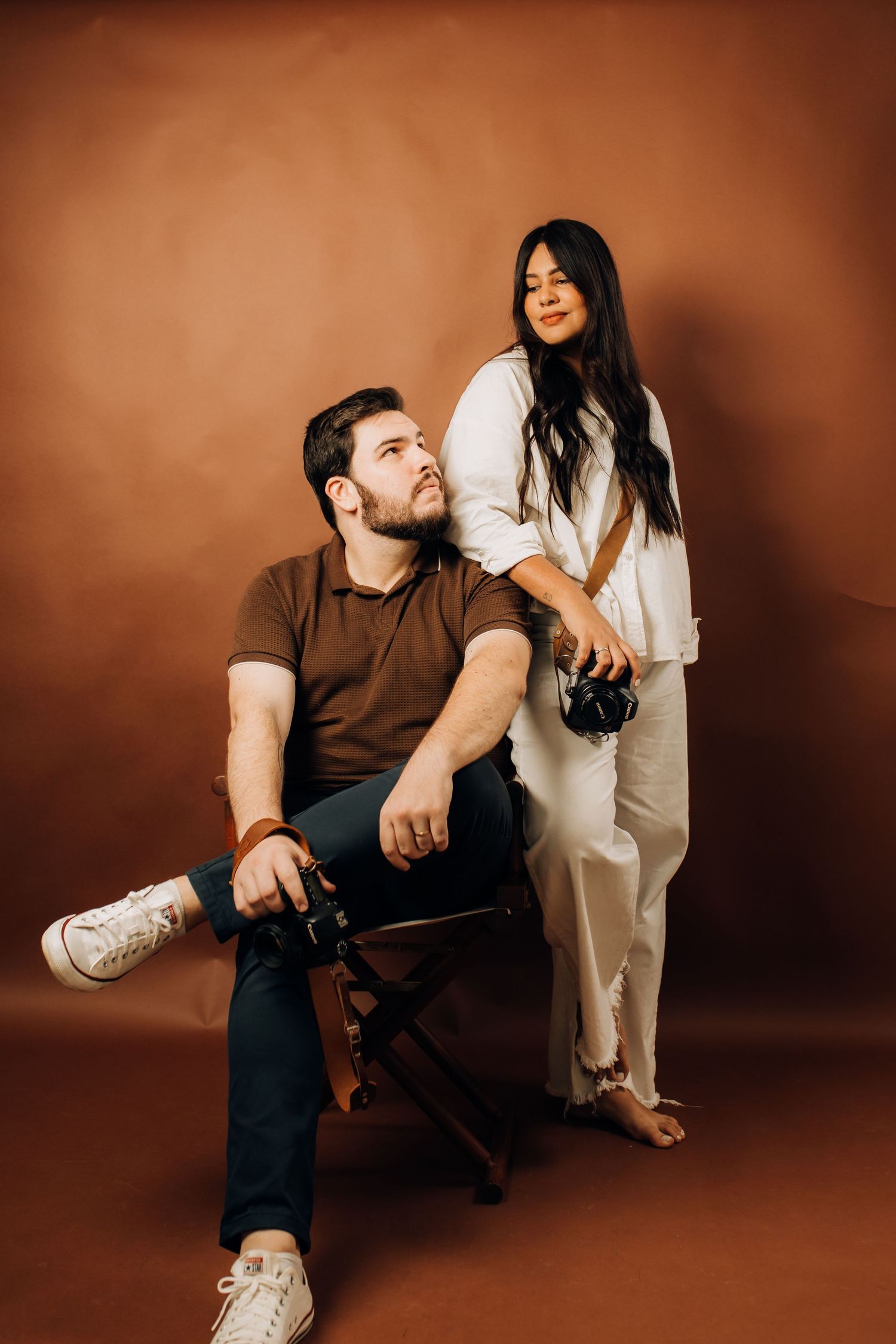 A man and woman pose in a studio with brown backdrop, both holding cameras, wearing casual outfits.