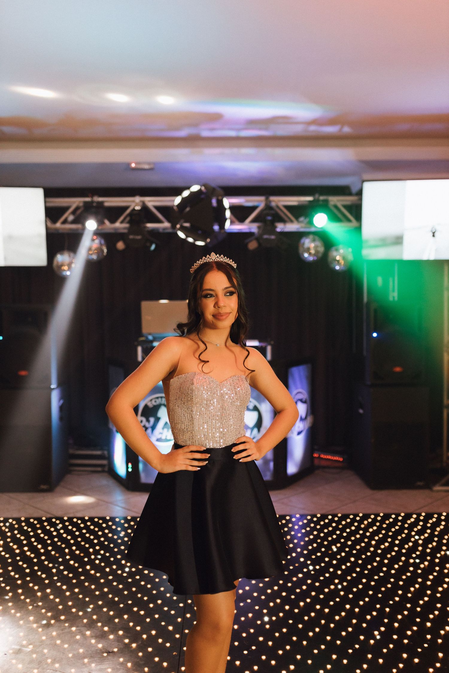 Young woman in silver top, black skirt, and tiara poses on LED dance floor at quinceañera party.