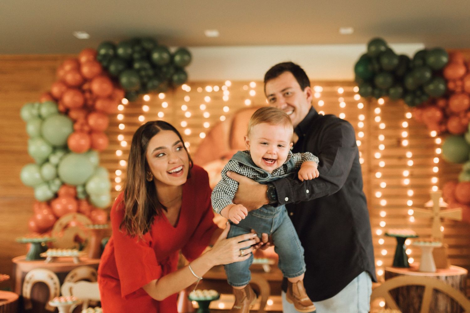 Happy family celebrating baby's first birthday with green and orange balloon decorations and string lights.