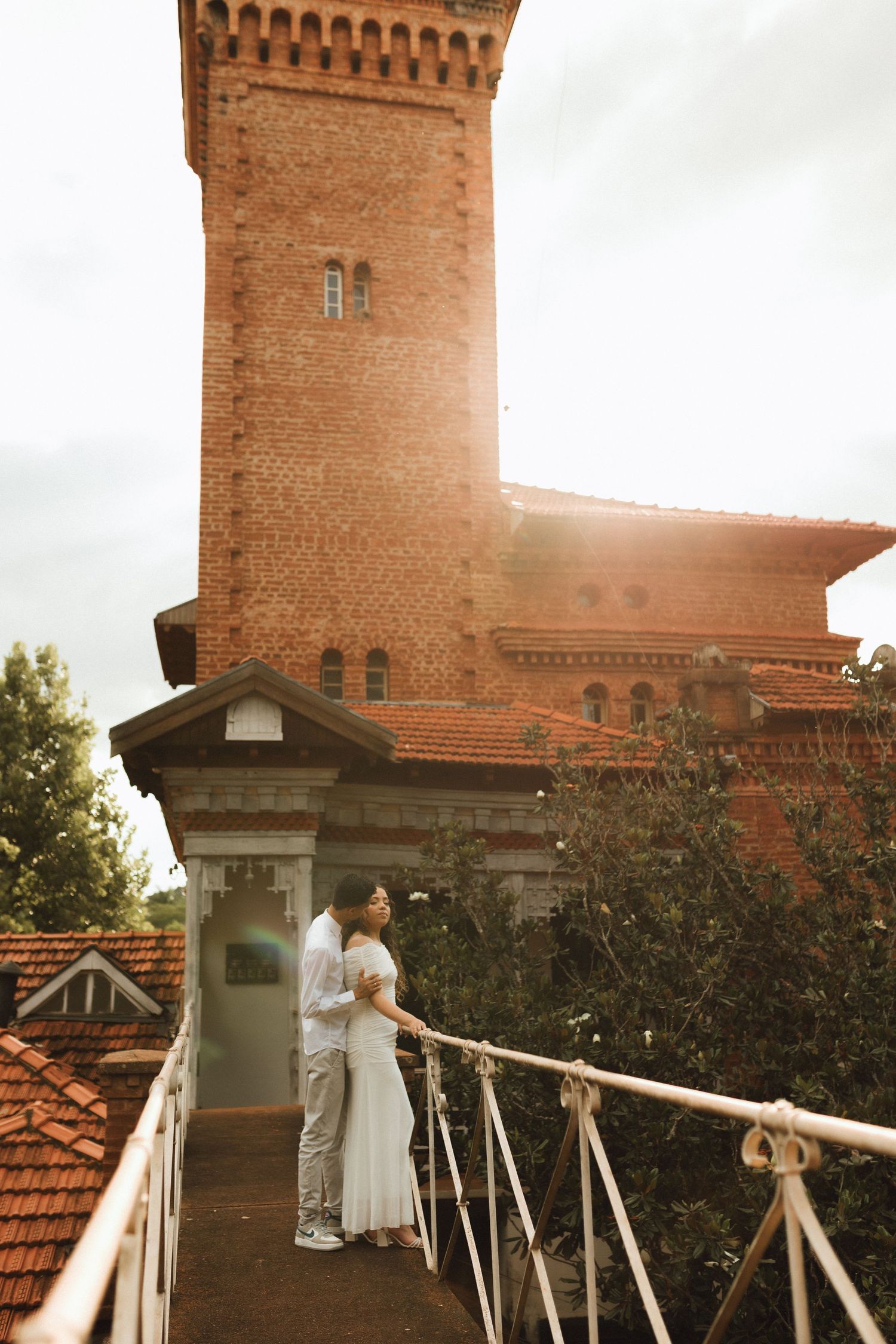 Romantic couple embracing on a bridge before a historic red brick tower in golden sunlight.