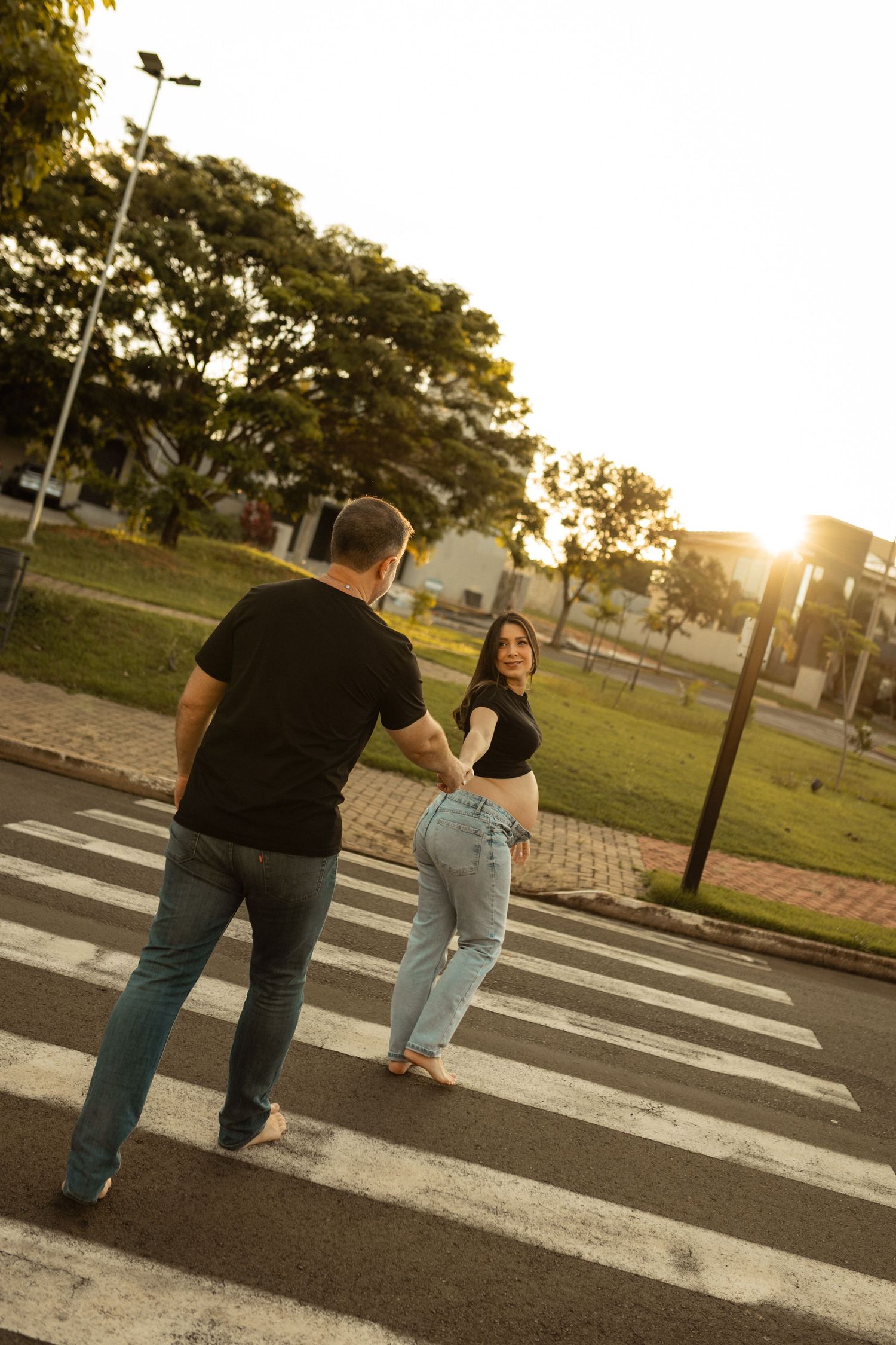 Pregnant couple holding hands while crossing a zebra crossing at golden hour sunset in urban park.