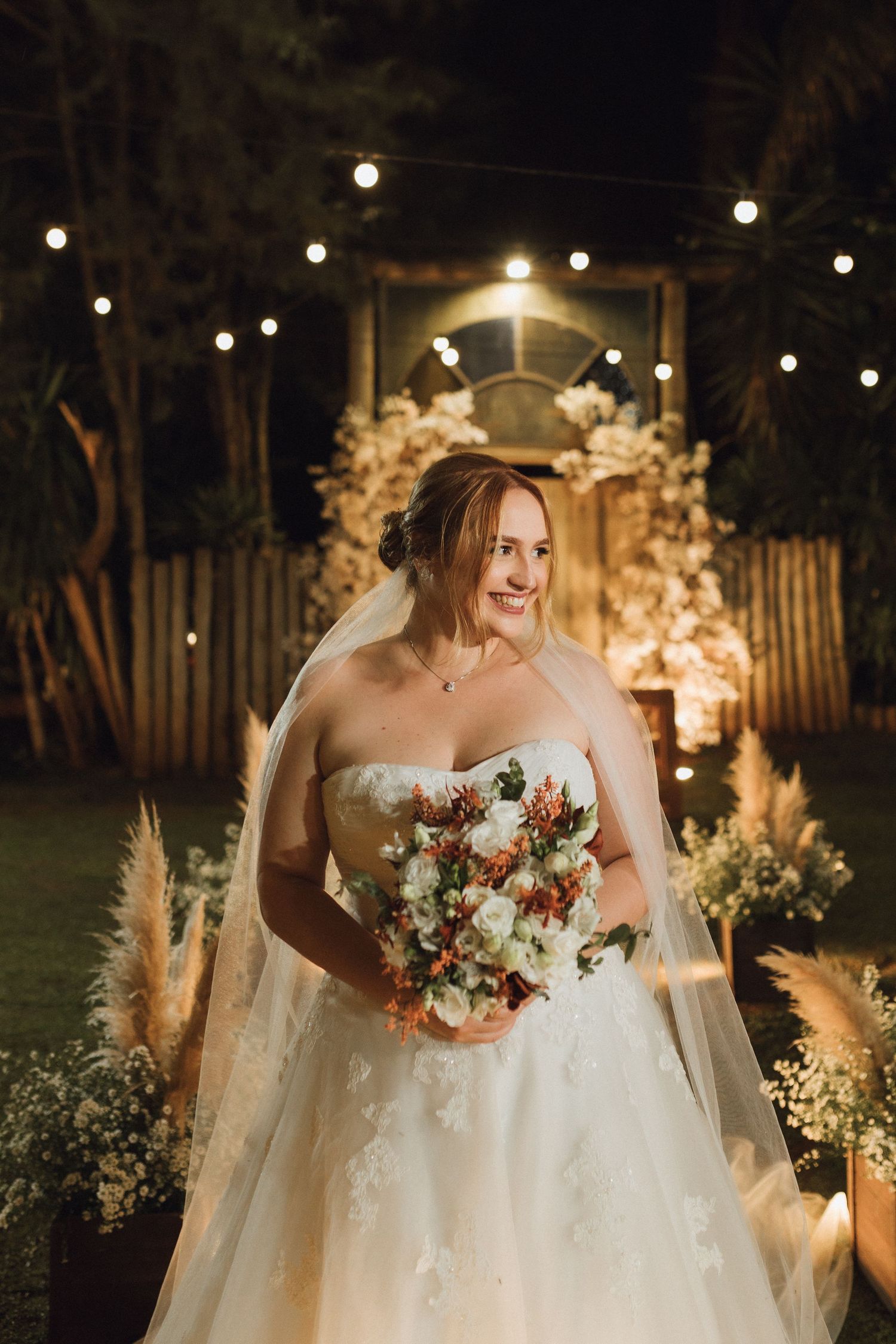 Radiant bride in strapless lace gown holding bouquet smiling before floral arch at night.
