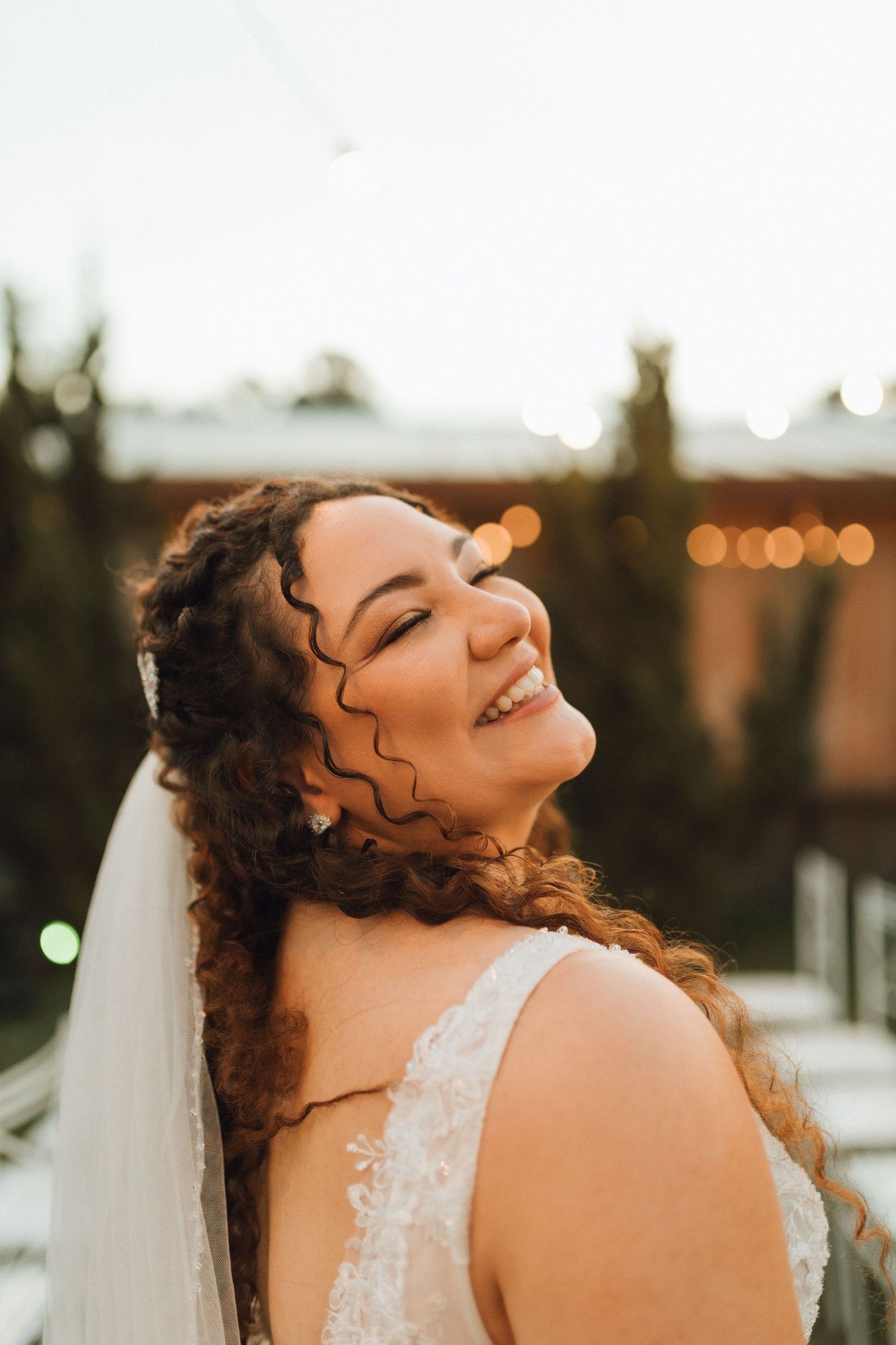 Radiant bride with curly hair and veil laughs joyfully in golden hour light at her outdoor wedding.