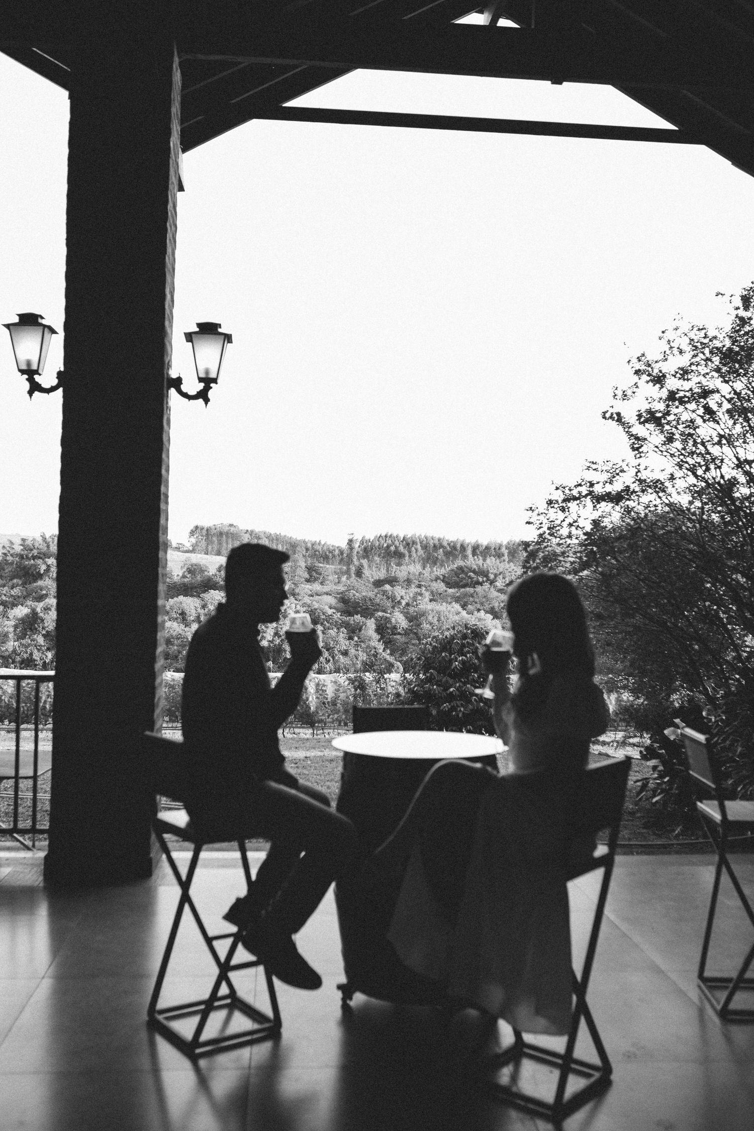 Black and white silhouette of a couple sitting at a table on a terrace, sharing drinks with a scenic landscape backdrop.