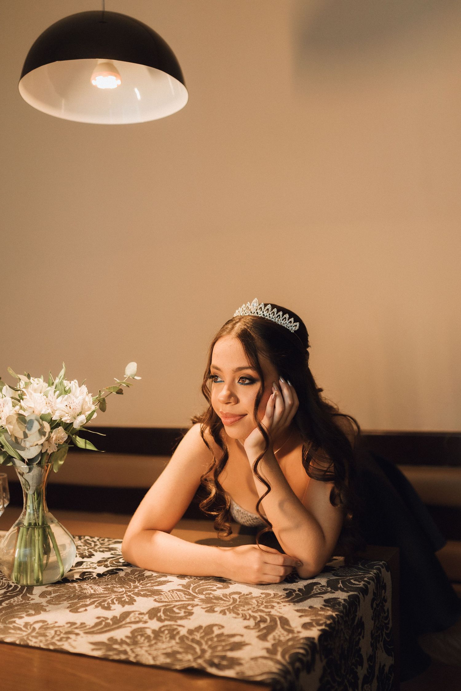 Young woman in black dress and diamond tiara rests at decorated table with white flowers in warm light.