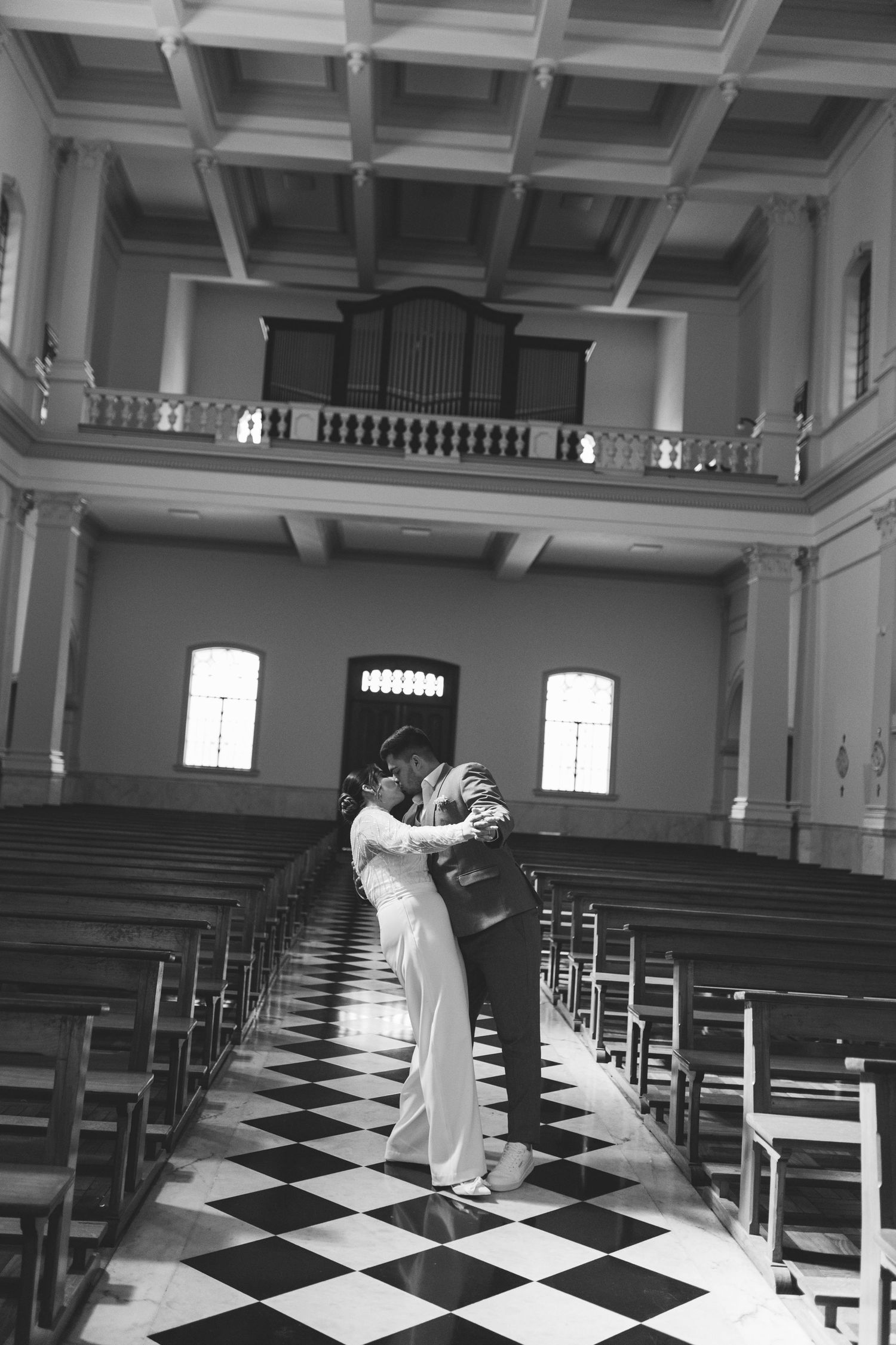 Black and white photo of a couple embracing in a church aisle with checkered floors and wooden pews.