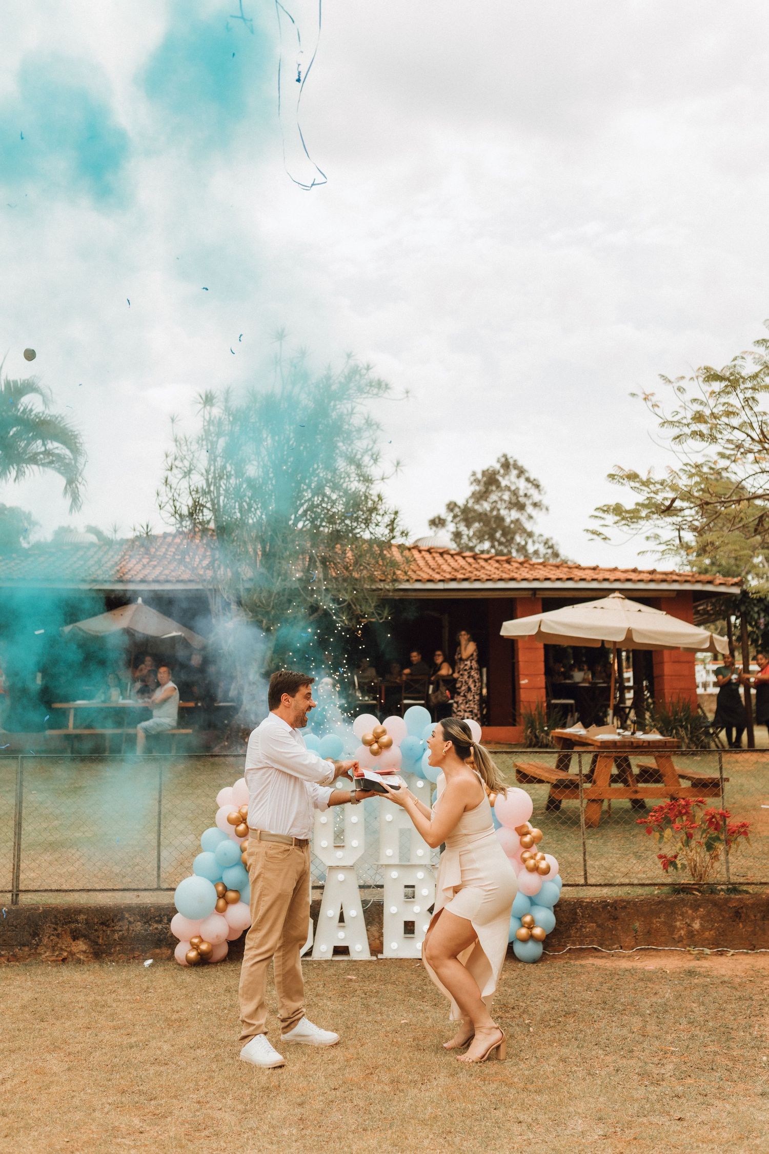 Excited couple celebrating blue smoke gender reveal for baby boy outdoors with balloon arch and OH BABY sign backdrop.