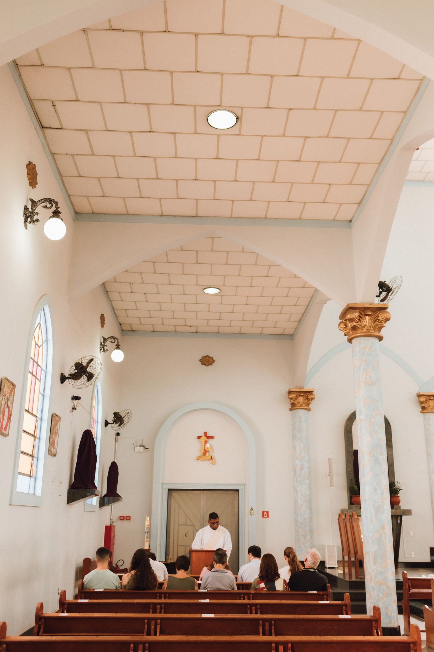 Interior of a small Catholic church with wooden pews, congregation seated, and a priest at the altar.