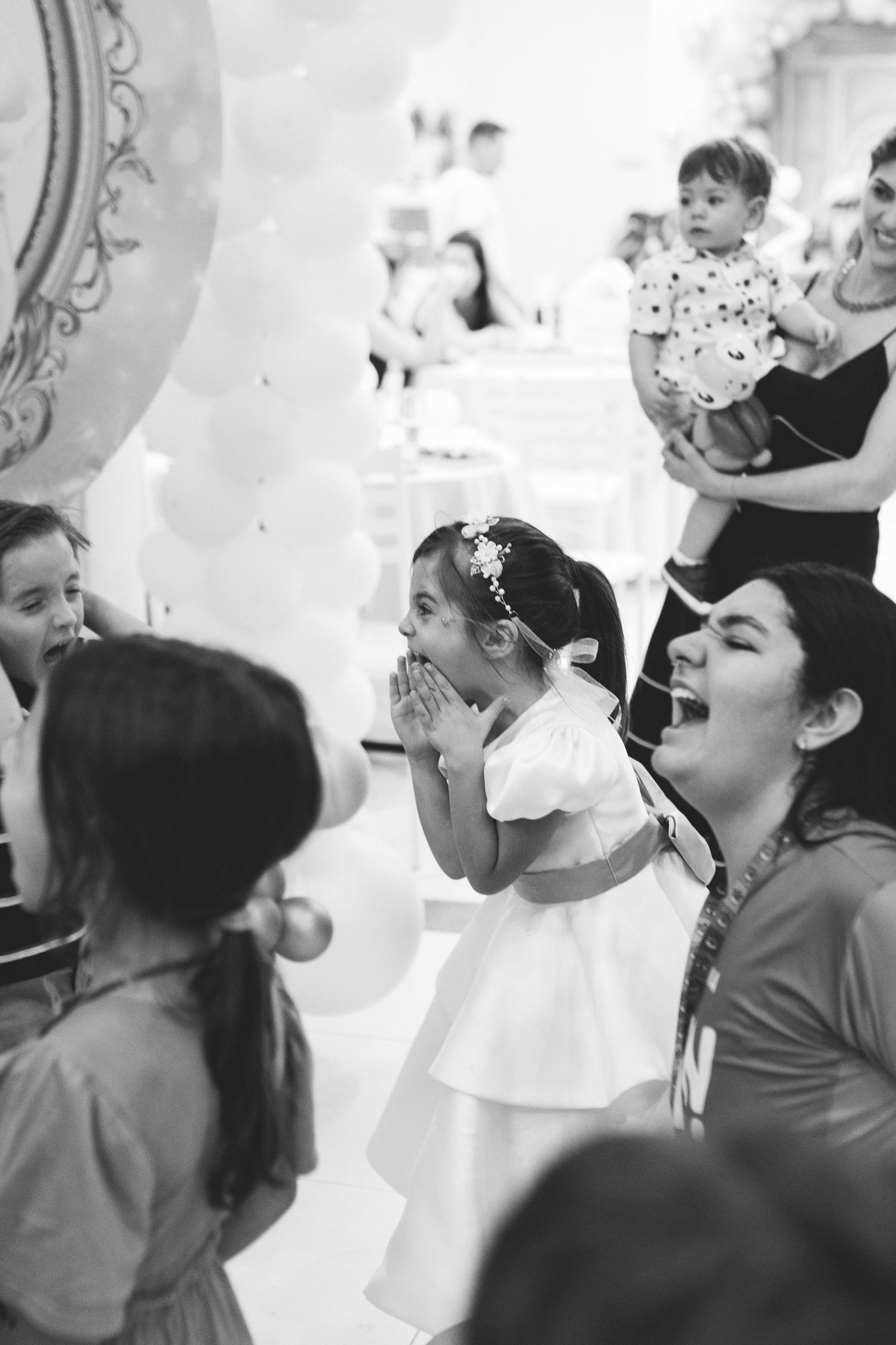 Black and white photo of excited children at a birthday party, girl in white dress with hands clasped in delight.