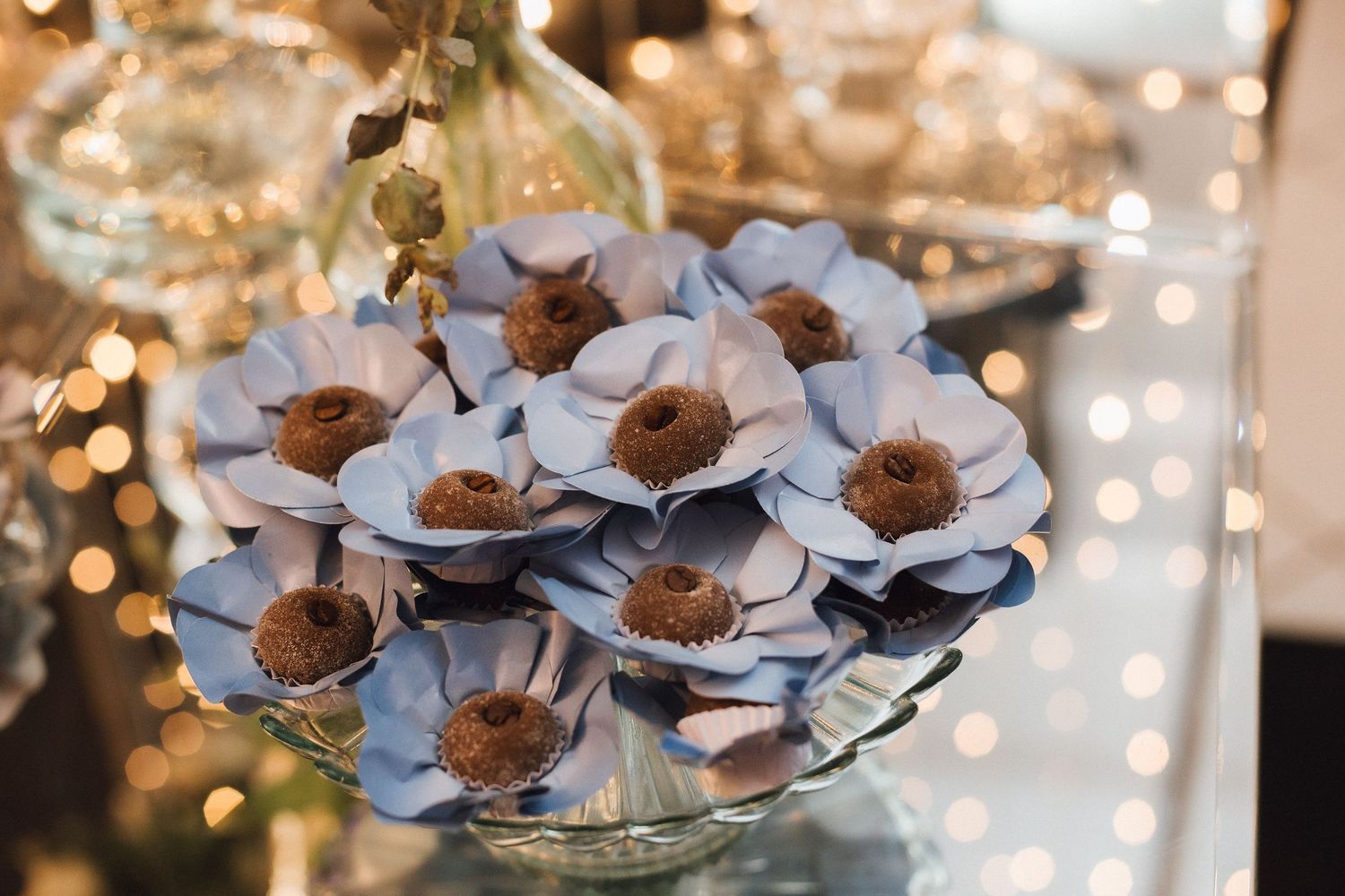 Decorative blue paper flower chocolates arranged in glass bowl with warm bokeh lights background.