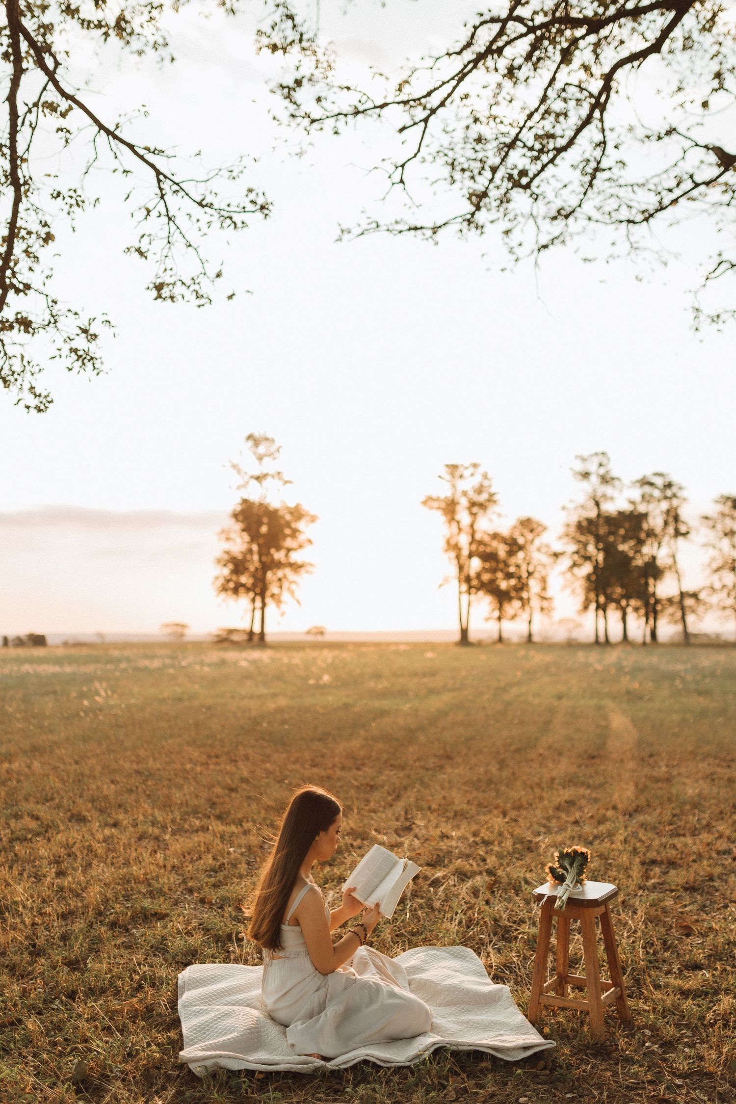 Young woman in white dress reading book on blanket in golden field at sunset with trees in background.