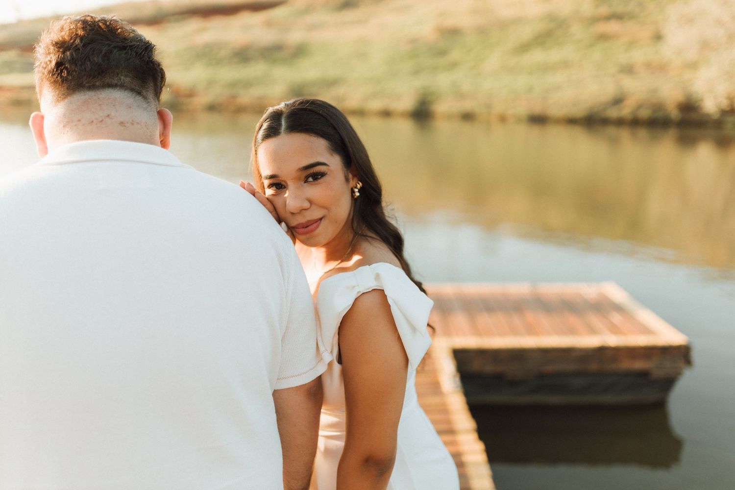 A woman in a white dress smiles over her shoulder near a lake dock during a romantic couples photo session.