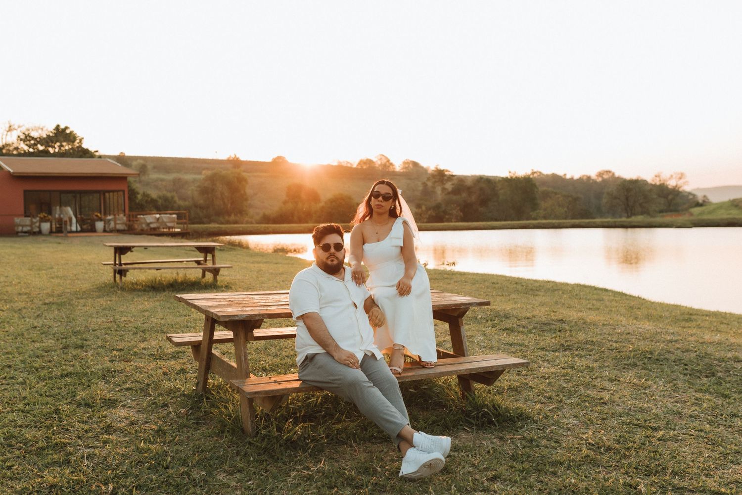 Couple in white outfits sitting at a wooden picnic table by a lake during golden hour sunset on green lawn.
