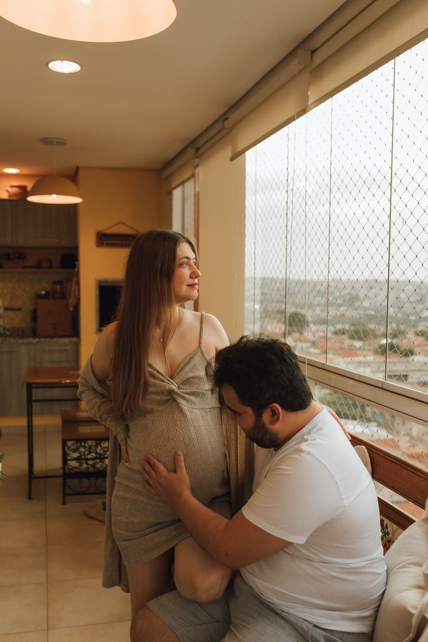 Pregnant woman gazes out window while partner tenderly kisses her baby bump in warm apartment.