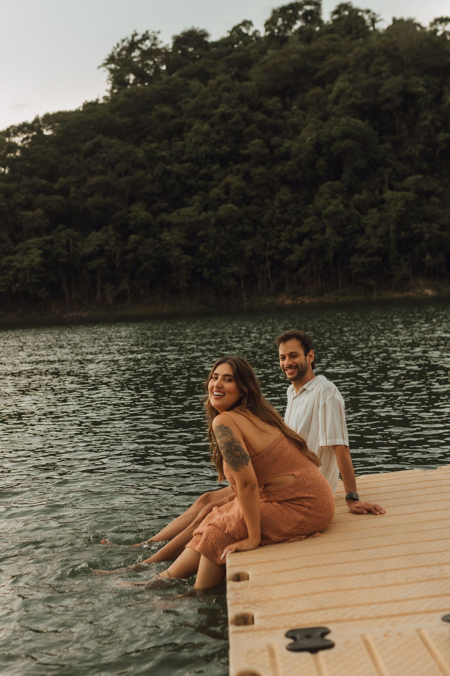 Couple sitting on wooden dock by lake surrounded by lush green forest.